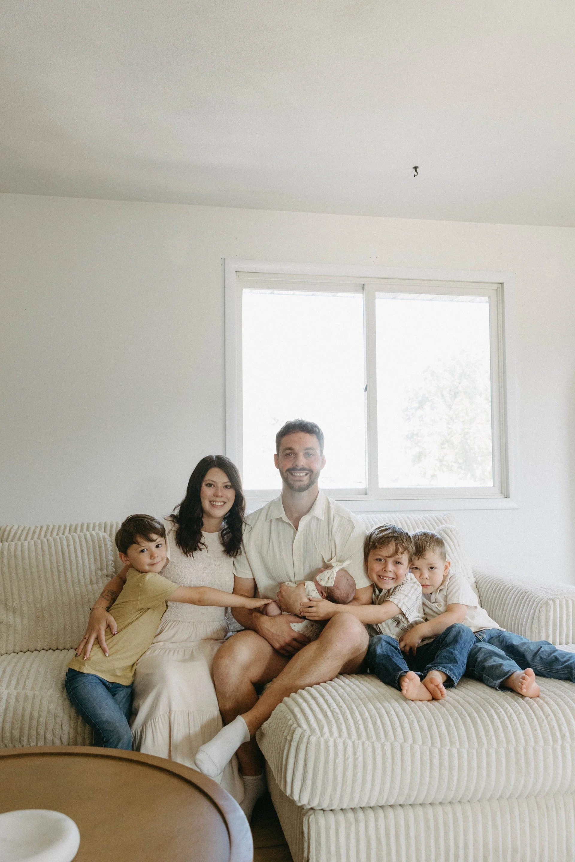 A family of seven sitting on a beige couch in a sunlit living room, with a man holding a newborn baby, surrounded by children, some touching the baby, all smiling, with a large window in the background.