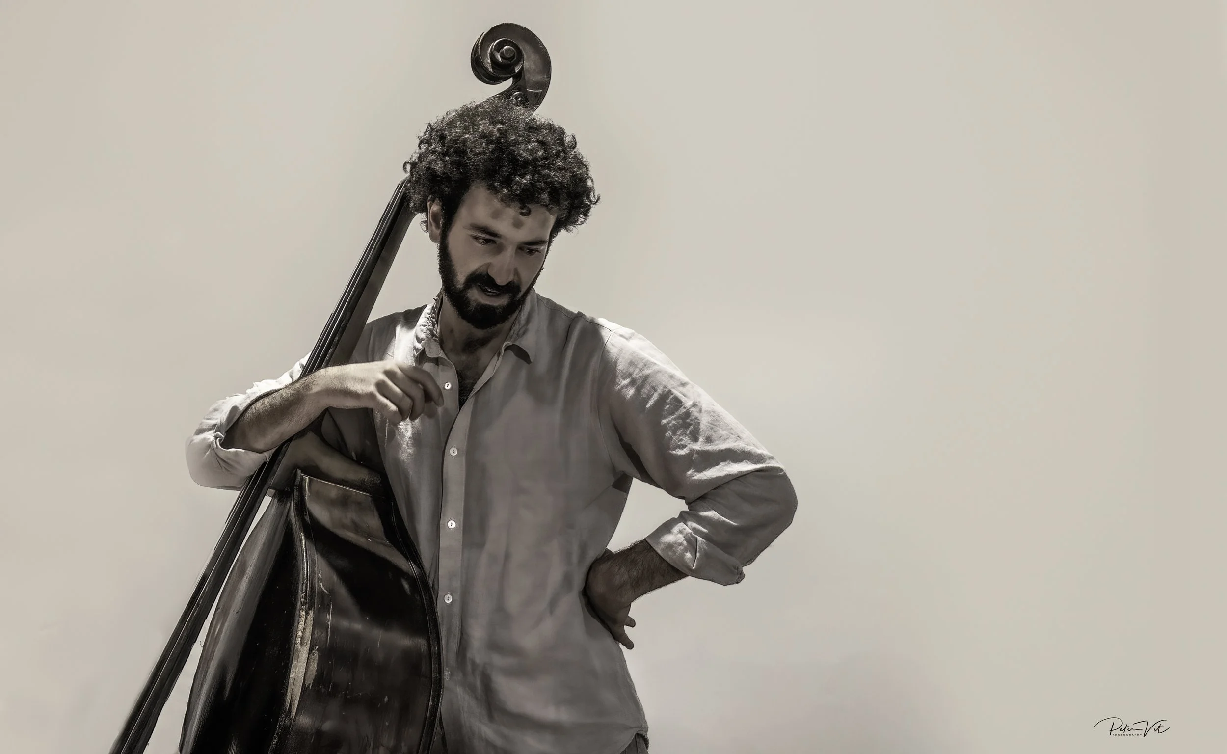A man with curly dark hair and a beard standing against a plain light-colored background, holding a contrabass with the neck resting over his shoulder, looking down with a thoughtful expression.