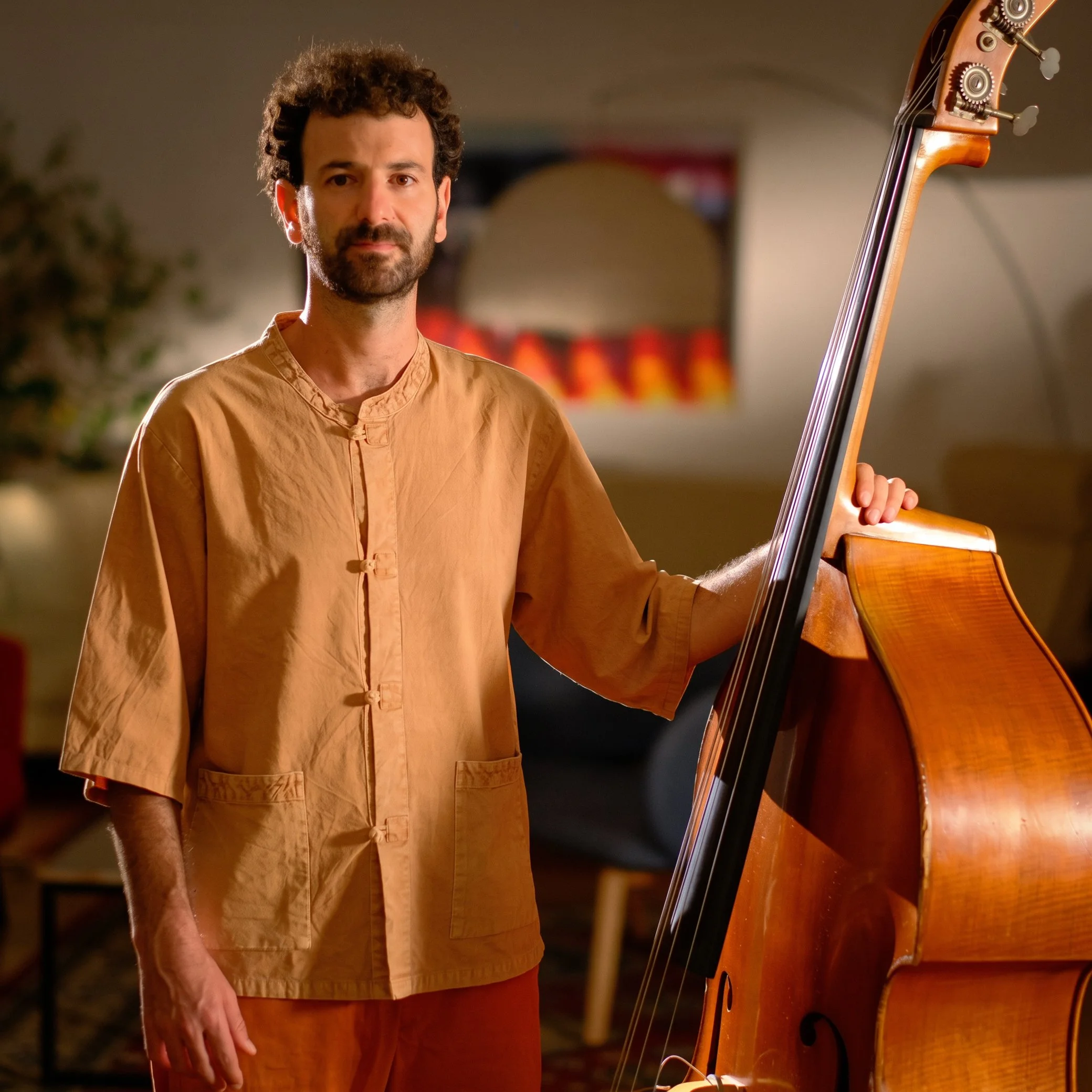 A man with curly hair, a beard, wearing a traditional orange Asian shirt, holding a wooden acoustic bass guitar in a warmly lit room with a blurred background.