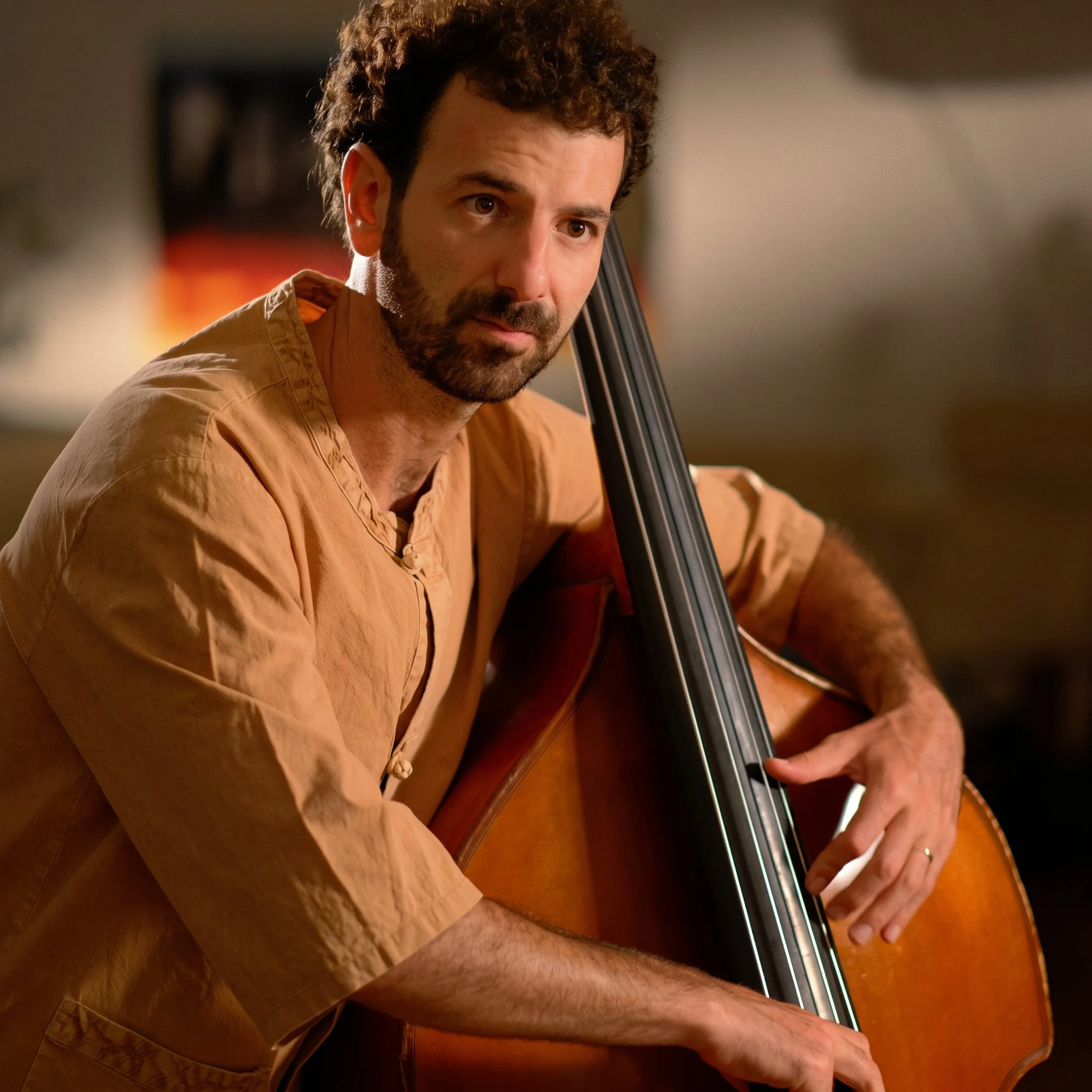 A man with curly brown hair and a beard playing the double bass, wearing a beige shirt, indoors with warm lighting.