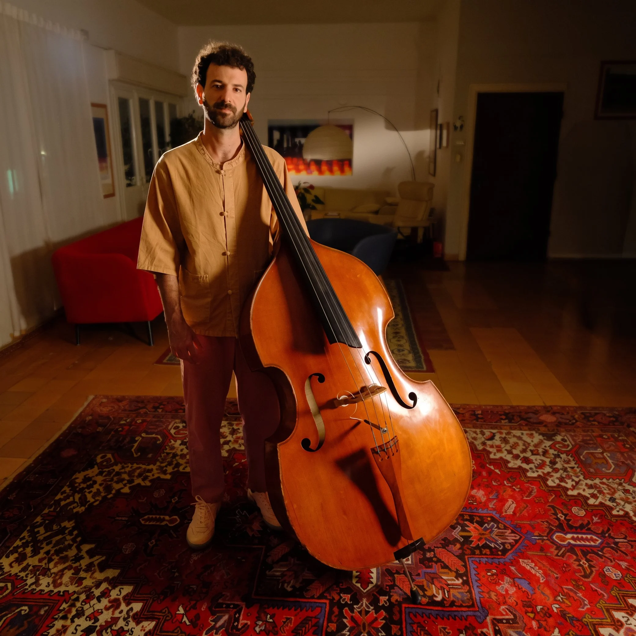 A man with dark curly hair and a beard standing indoors at night, holding a large upright double bass, wearing a tan traditional Asian-style shirt, in a warmly lit room with a red couch, patterned rugs, and modern art on the walls.