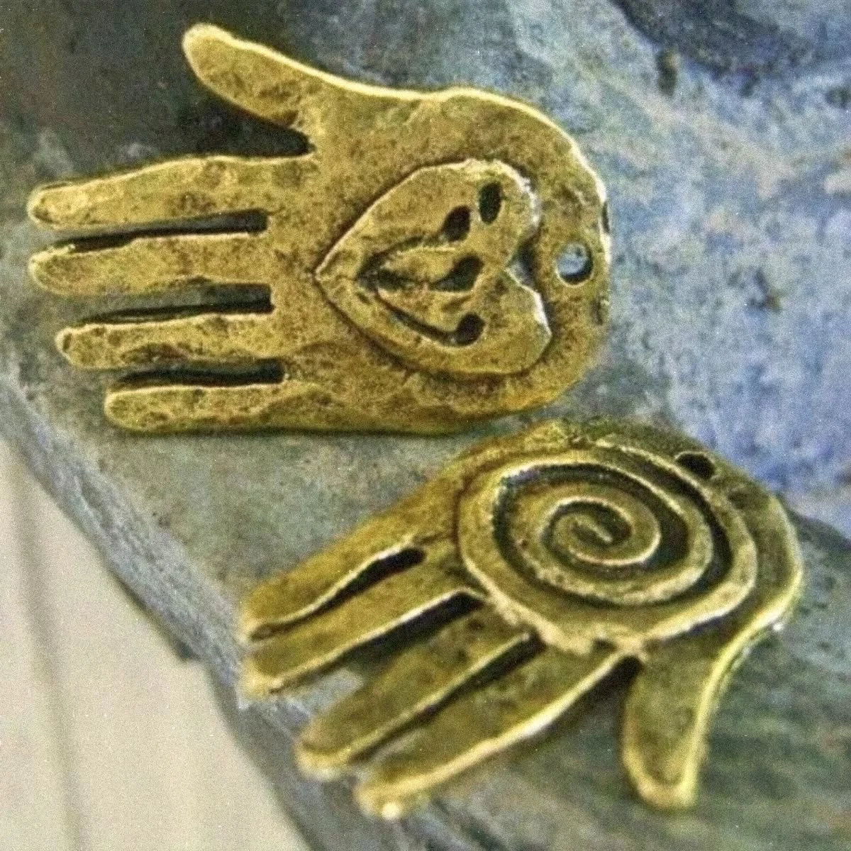 Close-up of two golden metal charms, each shaped like a hand with a face, one with spread fingers and the other with a spiral at the wrist.