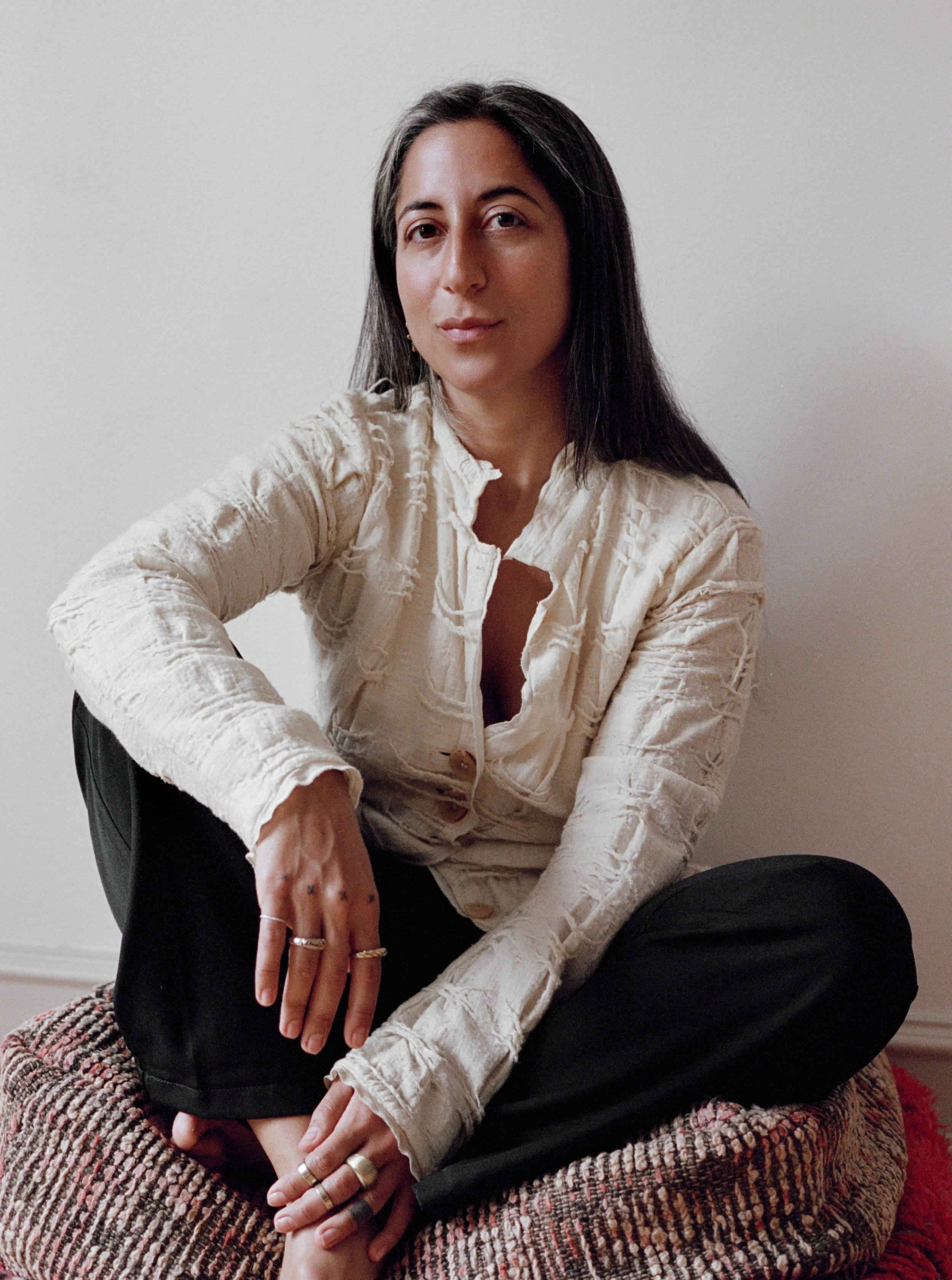 A woman with long dark hair and light skin sitting on a multicolored textured round cushion in front of a plain white wall. She is wearing a cream-colored, textured long-sleeve shirt and black pants. She has rings on her fingers and is sitting in a relaxed pose with her left hand resting on her knee and her right hand hanging in front of her.