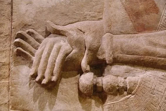 Close-up of ancient stone carving showing two hands, one holding a scepter or staff, with intricate patterns on the garment.