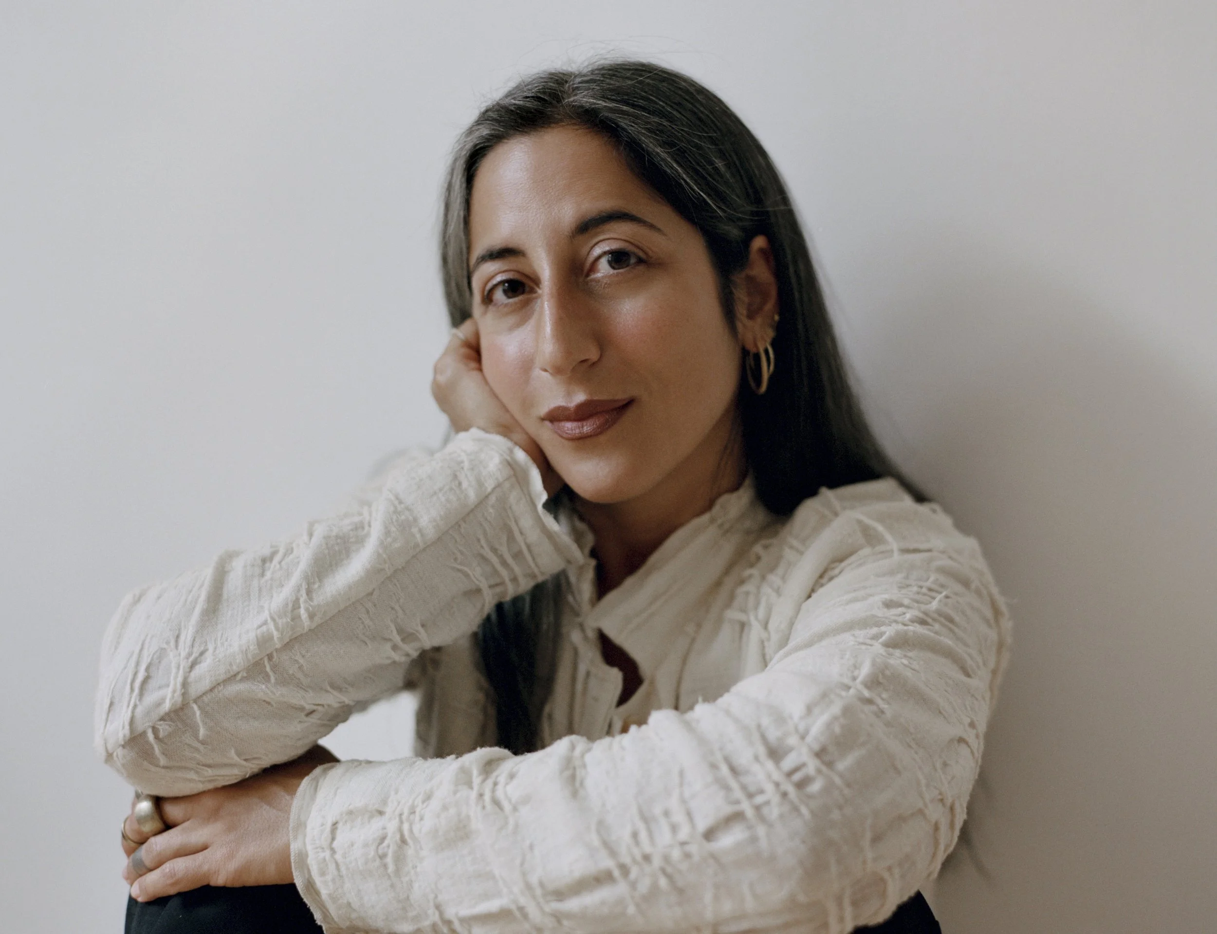 A woman with dark hair and hoop earrings resting her head on her hand, wearing a cream textured sweater, against a plain light background.