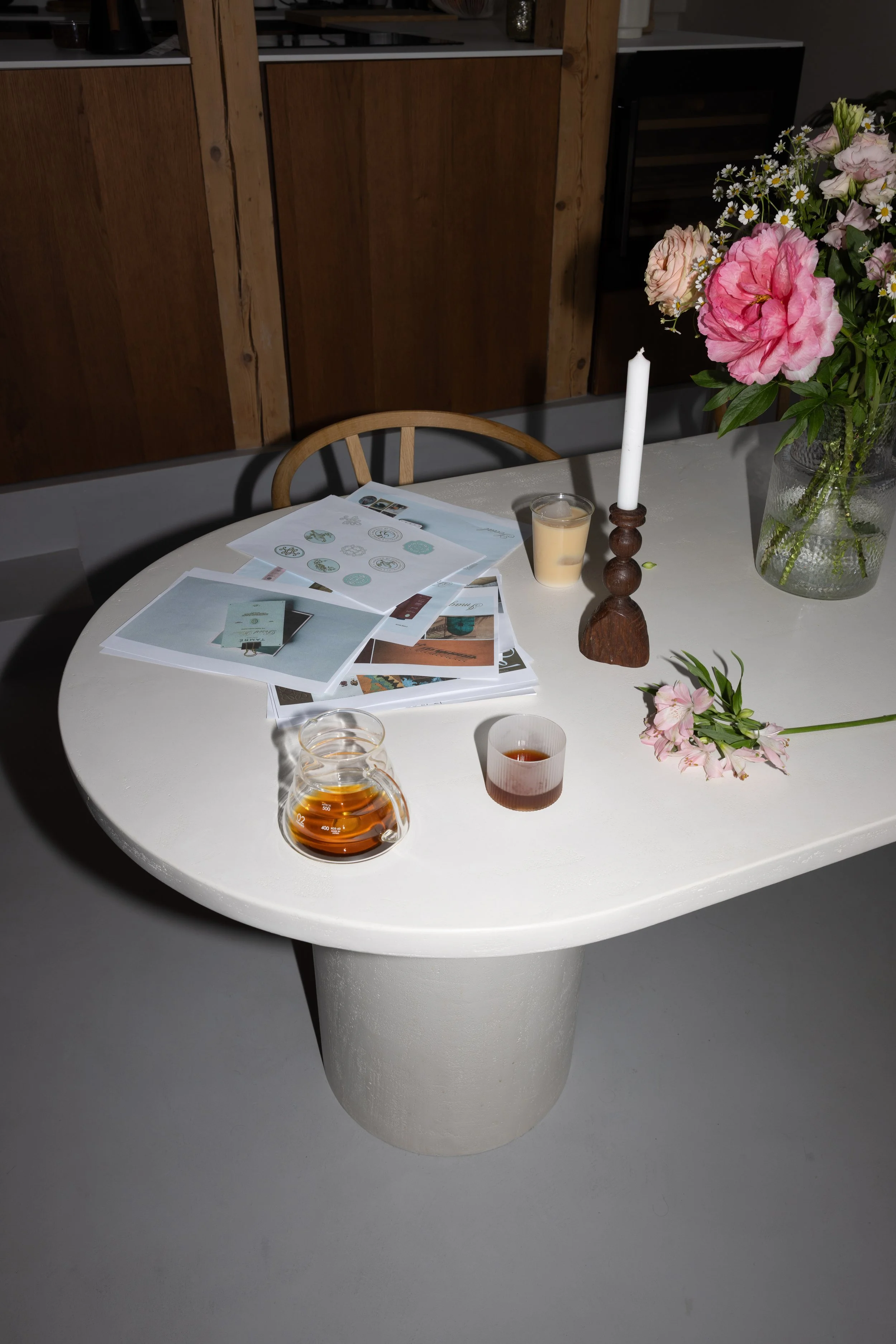 A white round table with a floral arrangement in a vase, a candle, and various papers with printed images and text. There are drinking glasses with beverages and a wooden chair nearby. The background features a wooden cabinet and a black appliance.