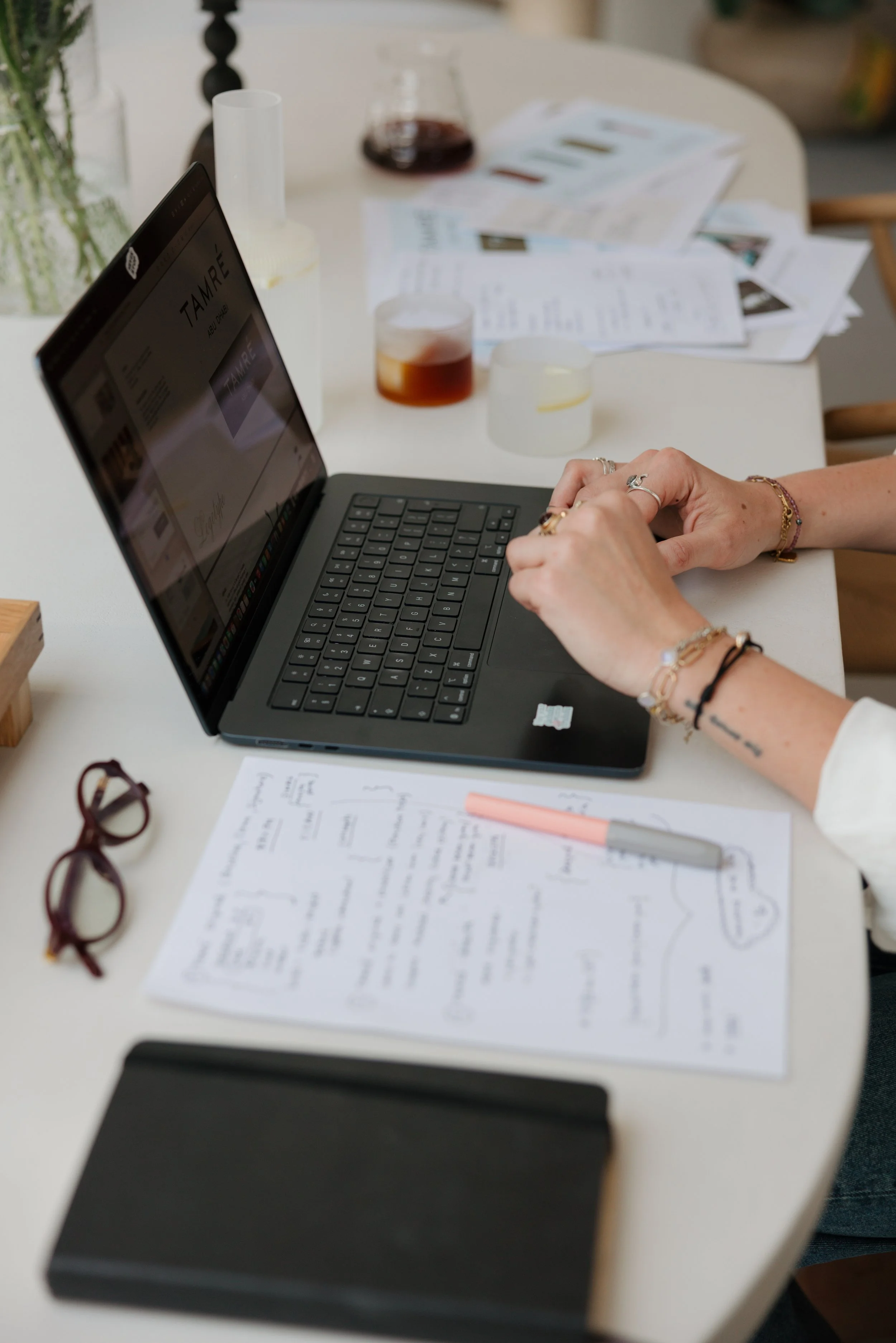A desk with a laptop, glasses, papers, and drinks, with hands resting on the laptop.