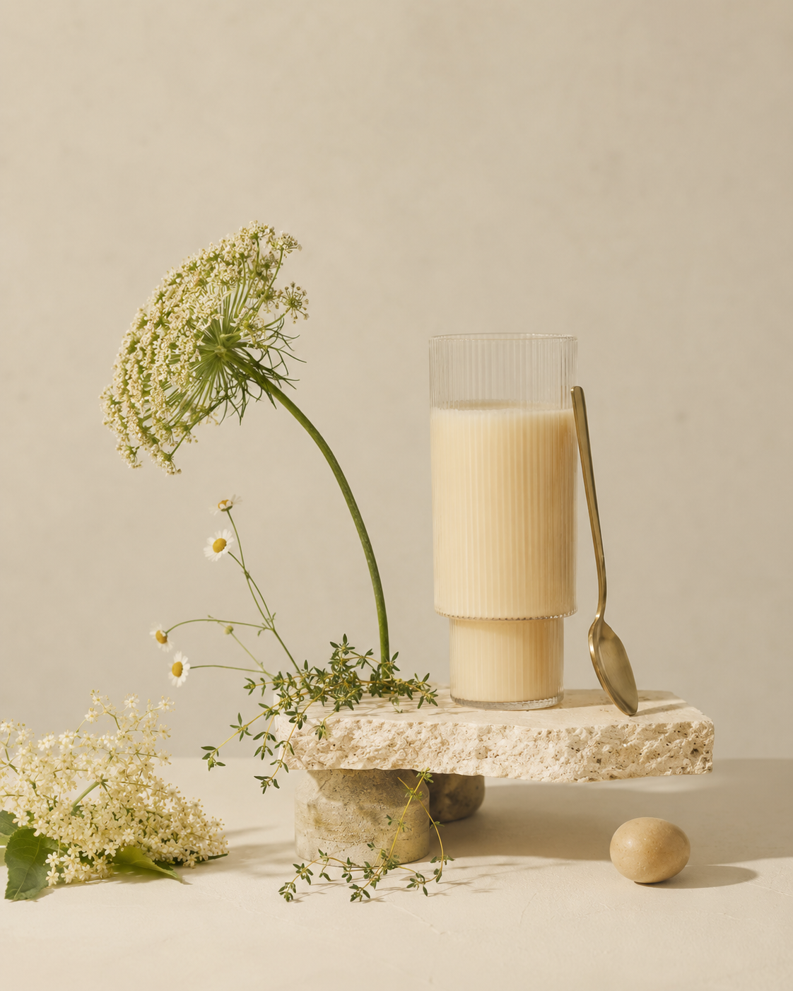 A glass of milk on a stone platform with a spoon, surrounded by white flowers and greenery against a plain background.