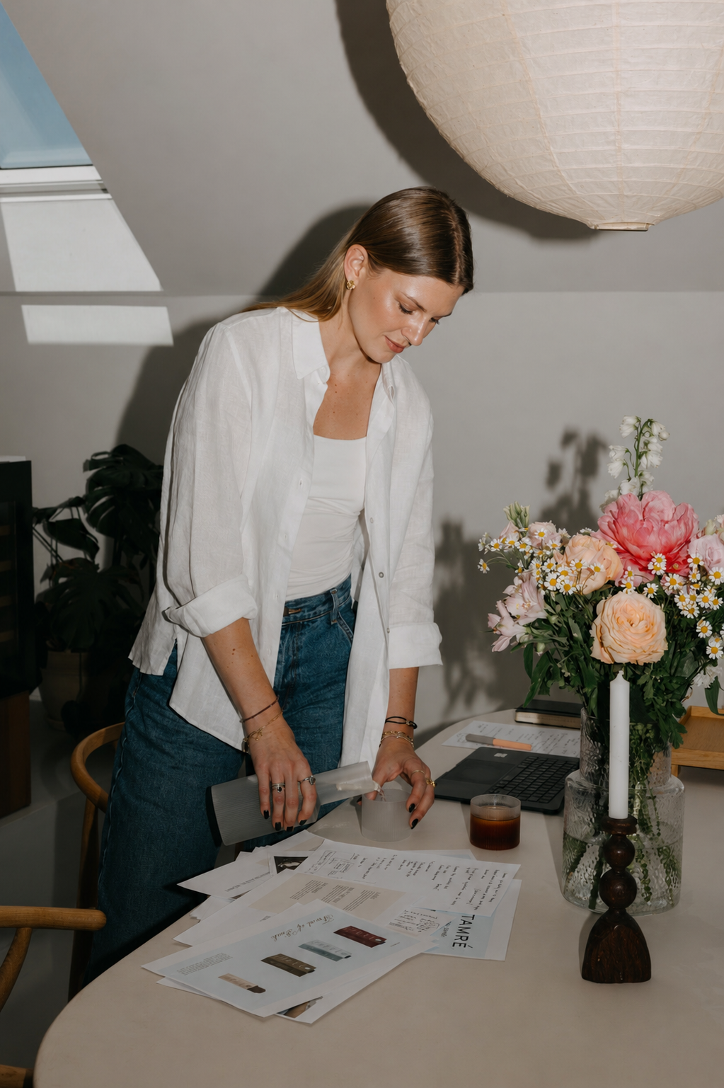 A woman in a white shirt and jeans is pouring a beverage into a glass on a table, with documents and a laptop, beside a vase of flowers and a candle.