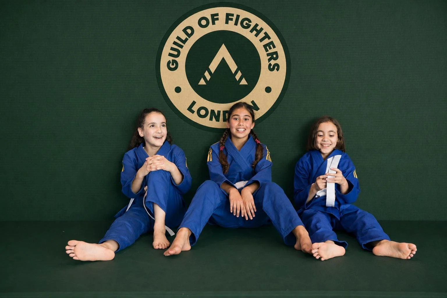 Three children's Brazilian Jiu Jitsu children in uniforms sitting on a green mat in front of a green wall with a logo reading 'Guild of Fighters London' and a stylized 'A' symbol.