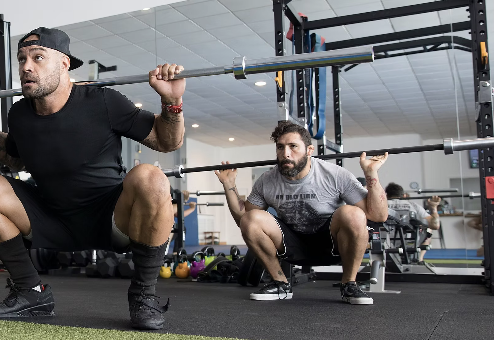 Two men squat with barbells on their shoulders in a gym in Wandsworth SW18. one man is in the foreground with a black cap worn backward, and the other man with a beard and tattoos, holding the barbell behind his head.