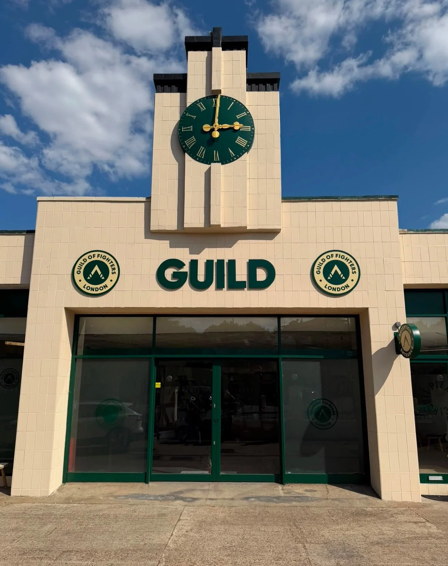 A building with a clock showing 3:00, with the word 'GUILD' and two circular logos of the Guild of Fighters London on each side of the entrance. The entrance has green sliding doors. The sky is partly cloudy.