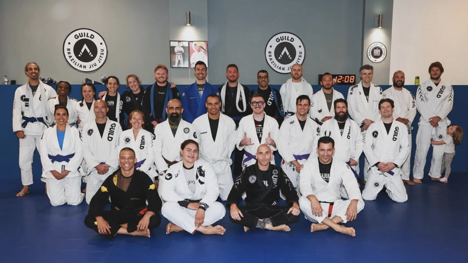 Group photo of Brazilian Jiu Jitsu practitioners in gi uniforms posing in a training facility with a sign that reads "Guild Brazilian Jiu Jitsu."