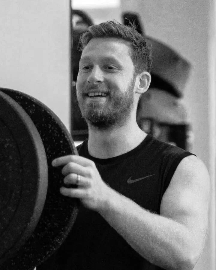A man with brown hair and a beard, smiling, wearing a black sleeveless Nike shirt, holding a weight plate in a gym.