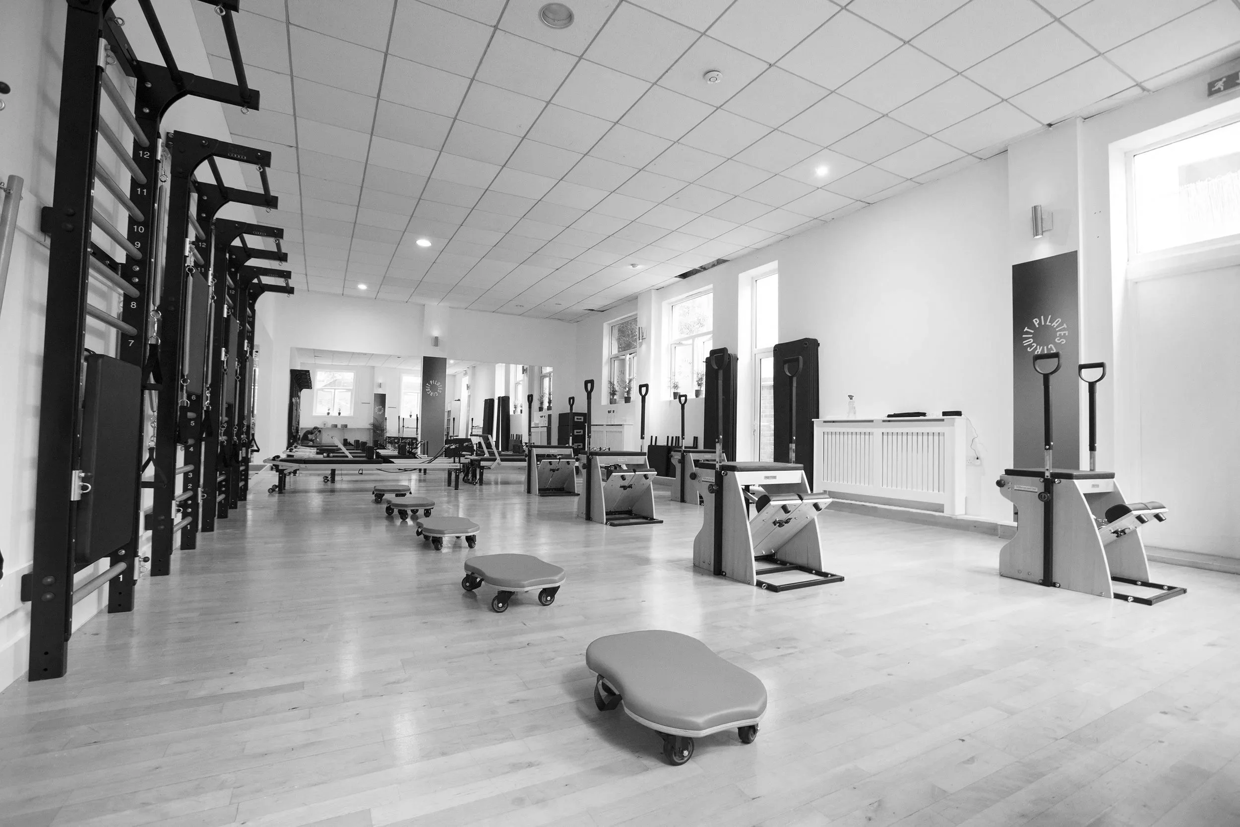 A spacious fitness room with wooden floors, white walls, and large windows. The room contains various gym equipment including squat racks, benches, exercise mats, and small rolling stools. The ceiling is tiled with recessed lighting. A pilates studio