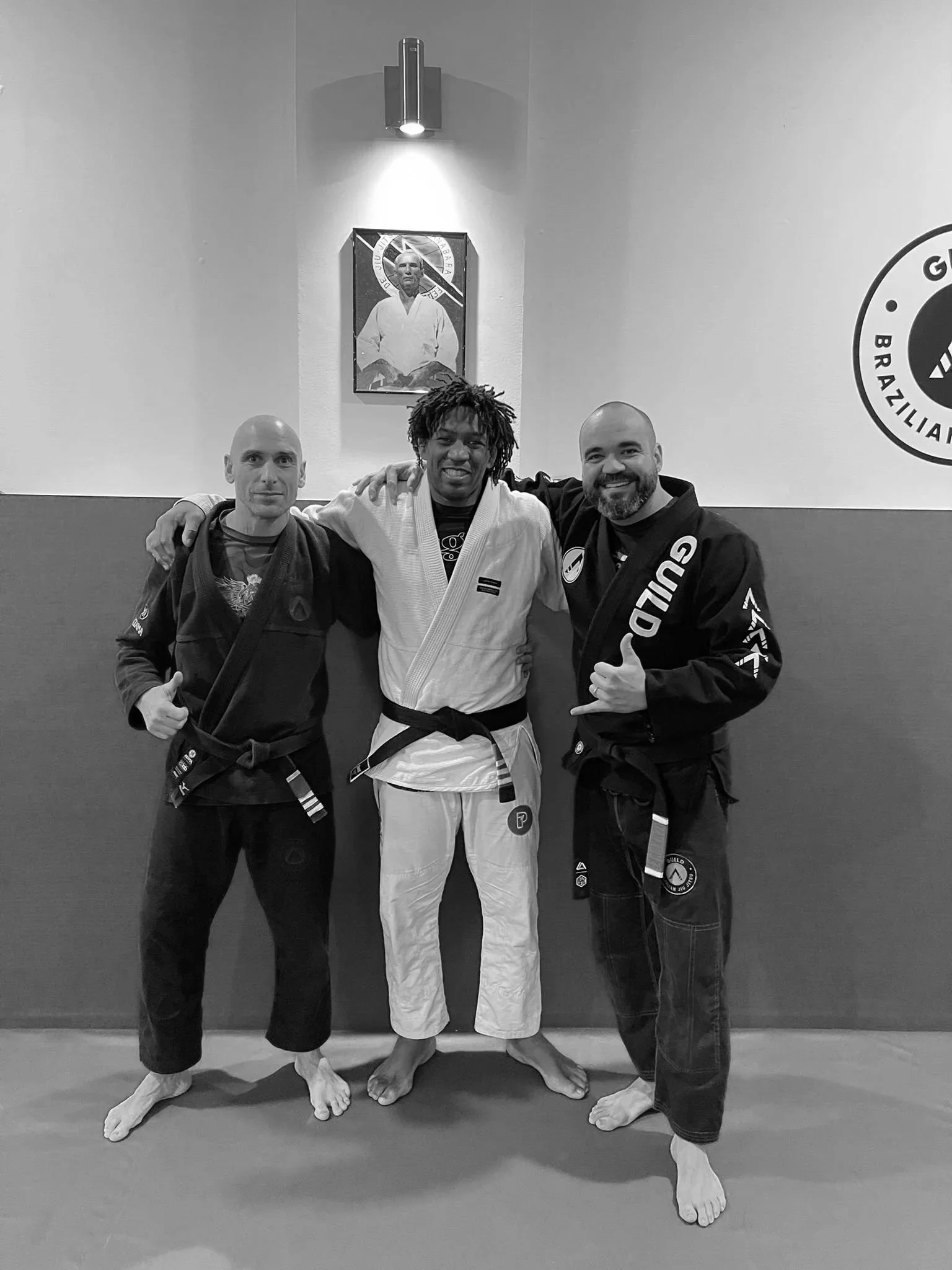 Three men in martial arts uniforms standing on a gym mat, posing with their arms around each other.