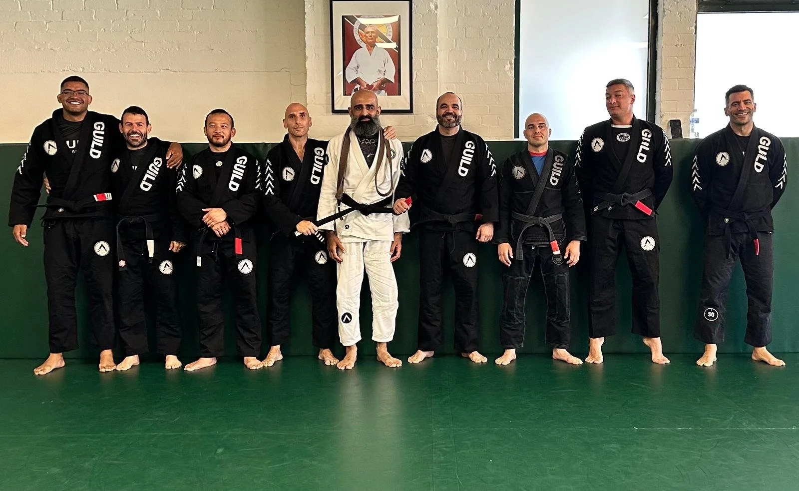 Group of martial arts practitioners in black gis standing on a green mat, with one instructor in a white gi, inside a dojo with a brick wall and a framed photo of a martial arts master in Wandsworth, London.