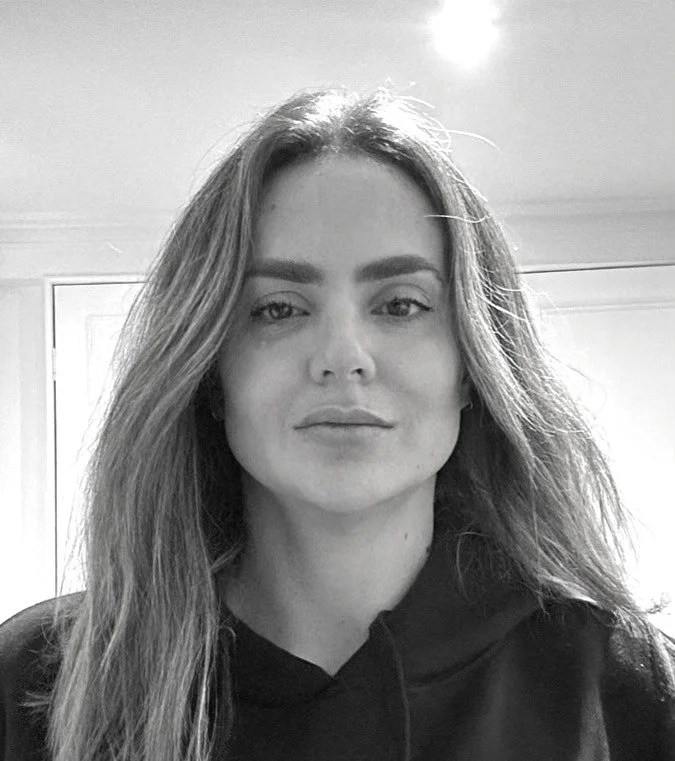Black and white photo of a woman with long, wavy hair, wearing a dark hoodie, looking directly at the camera with a neutral expression.