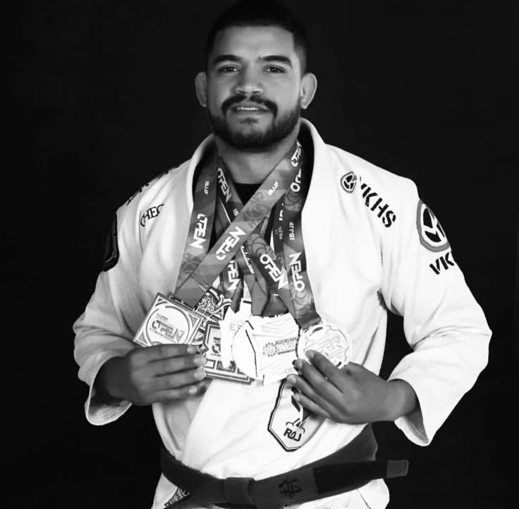 A jiu jitsu professor from guild of fighters BJJ academy in Wandsworth in a martial arts uniform with several medals around his neck, holding some certificates, standing against a dark background.