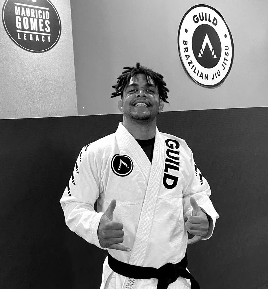 A BJJ instructor wearing a Brazilian Jiu-Jitsu gi with the word 'GUILD' on it, standing and smiling in a martial arts gym. The gym's wall features two circular logos: one saying 'Mauricio Gomes Legacy' and the other 'GUILD Brazilian Jiu Jitsu'.