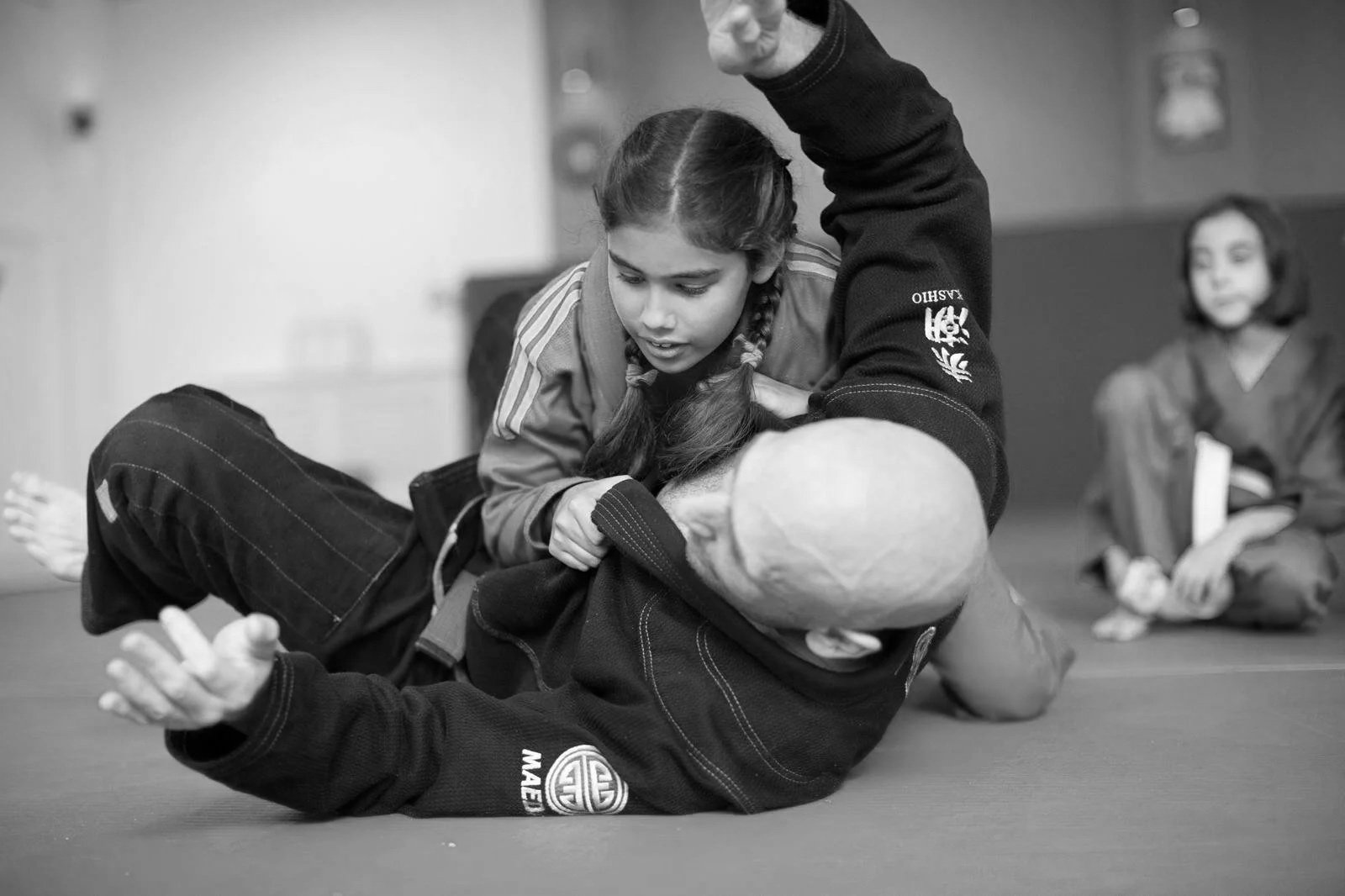 Two girls practicing Brazilian Jiu-Jitsu, one girl in a black gi on her back on the mat, another girl in a jacket and striped pants on top, demonstrating a grappling technique, with a third girl in a gi sitting in the background.