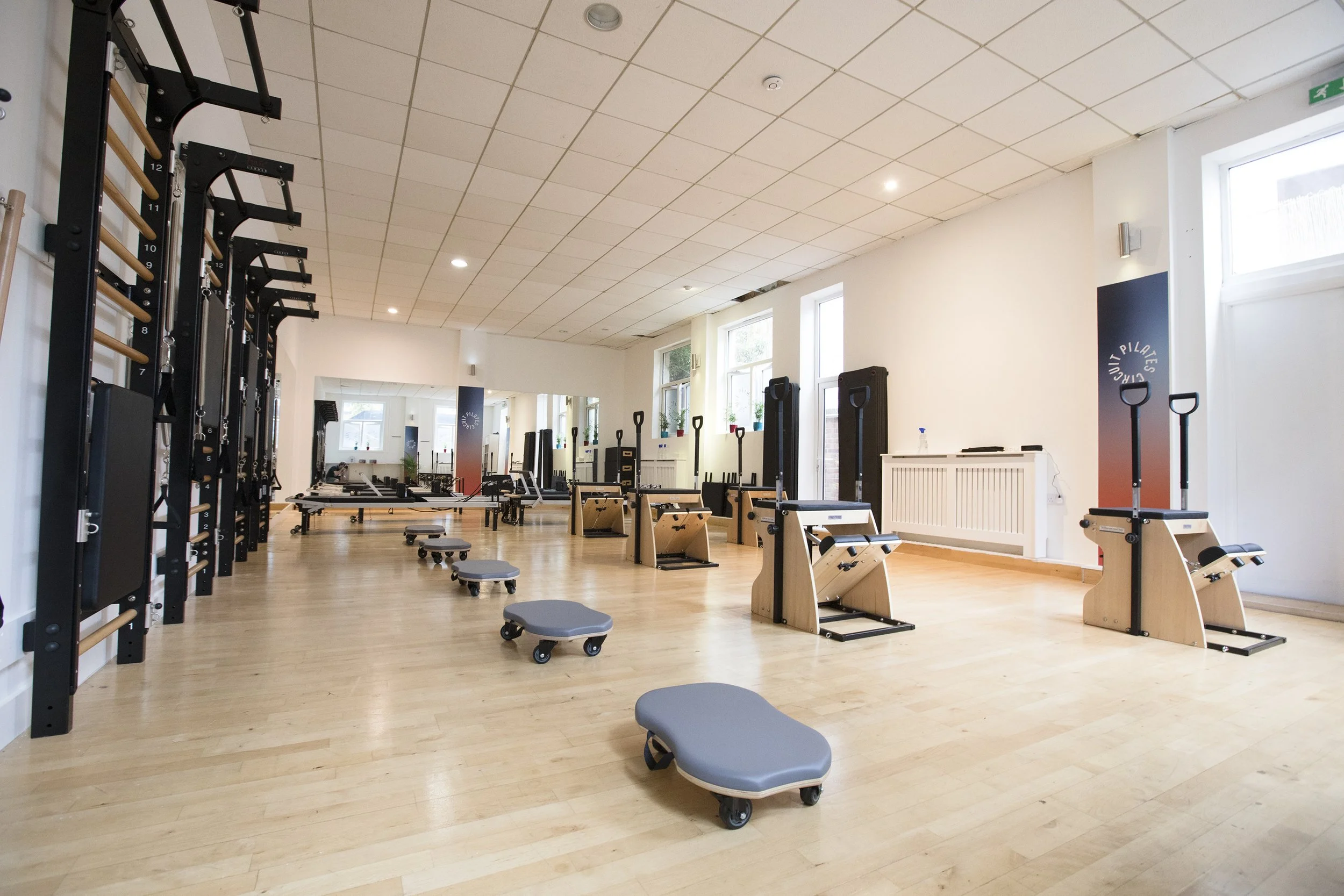 A spacious fitness room with wooden floors, white walls, and large windows. The room contains various gym equipment including squat racks, benches, exercise mats, and small rolling stools. The ceiling is tiled with recessed lighting. A pilates studio