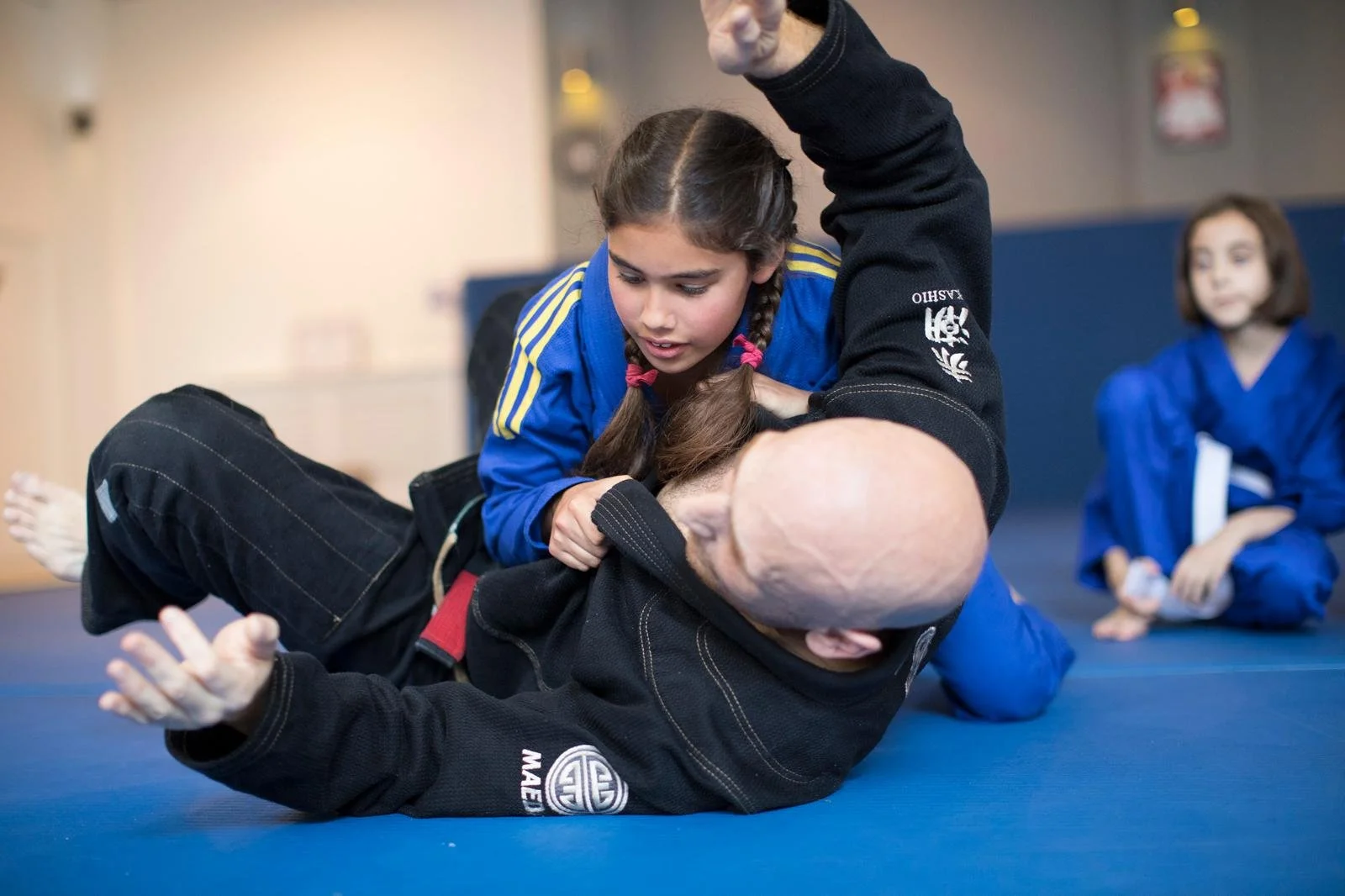 Young girl in a blue martial arts gi practicing Brazilian Jiu-Jitsu on a bald instructor on a blue mat, with another girl in a blue gi sitting in the background.