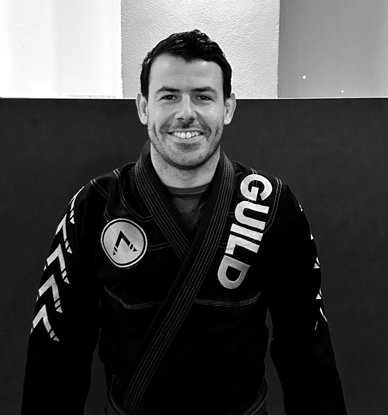 A jiu jitsu professor from guild of fighters Brazilian jiu jitsu academy in Wandsworth smiling in a Brazilian Jiu-Jitsu gi standing in front of a plain wall.