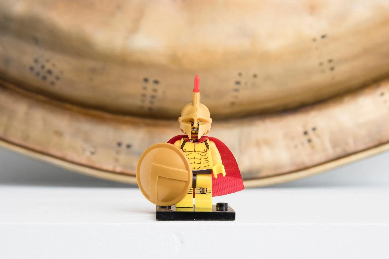 LEGO minifigure dressed as a Roman soldier with a red cape, shield, and helmet, standing on a black base in front of a large brass or bronze object.
