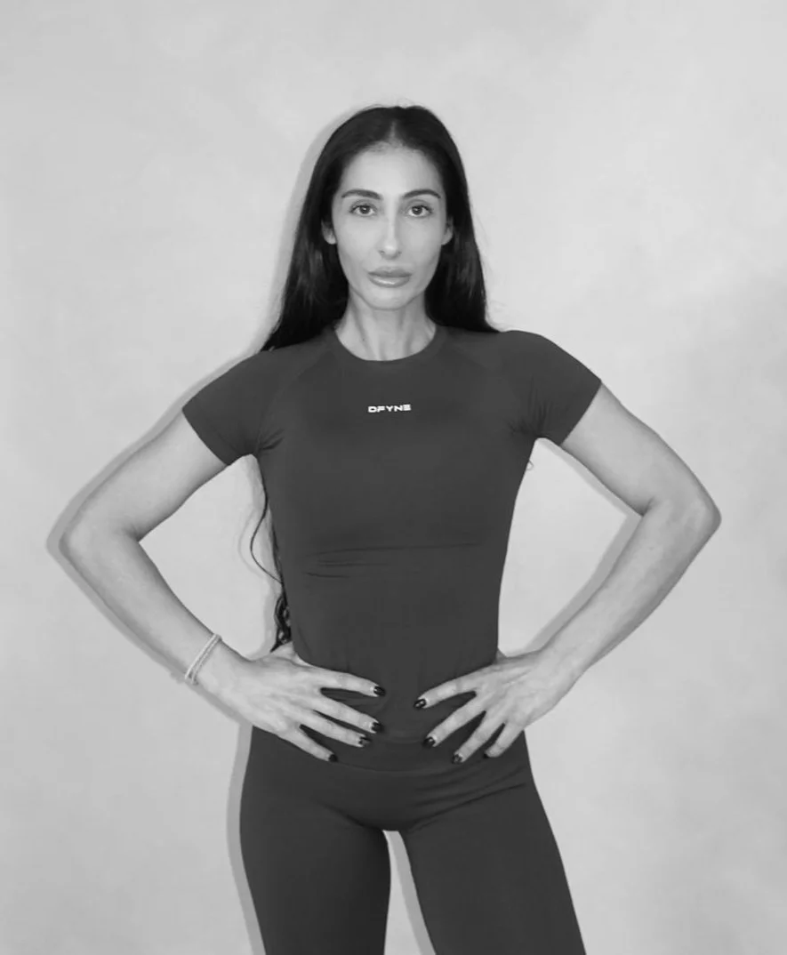 A woman with long dark hair wearing a fitted athletic t-shirt and leggings, standing with her hands on her hips against a plain wall. she is a post and pre natal fitness trainer in Wandsworth south west London.