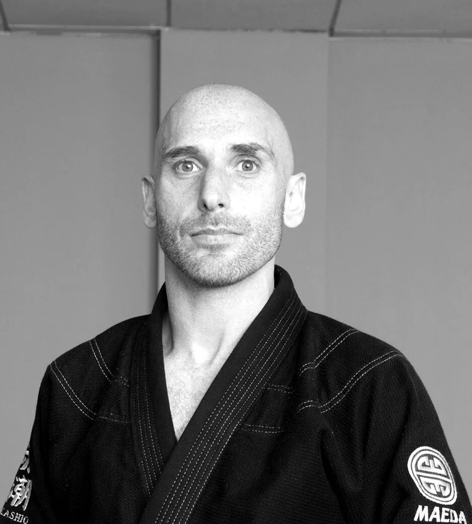 A man with a shaved head and beard wearing a black martial arts uniform with patches, standing indoors against a plain wall.