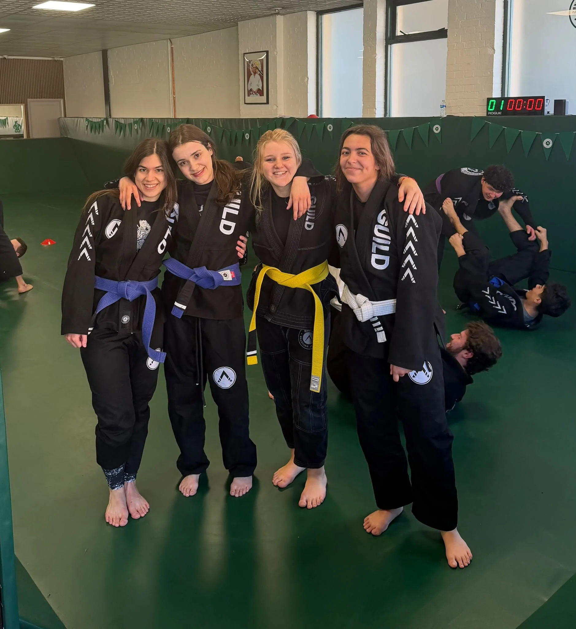 Four young teens Brazilian Jiu-Jitsu members in London with different colored belts pose together in a martial arts gym, smiling for the camera, with two young men practicing techniques on the mat in the background.