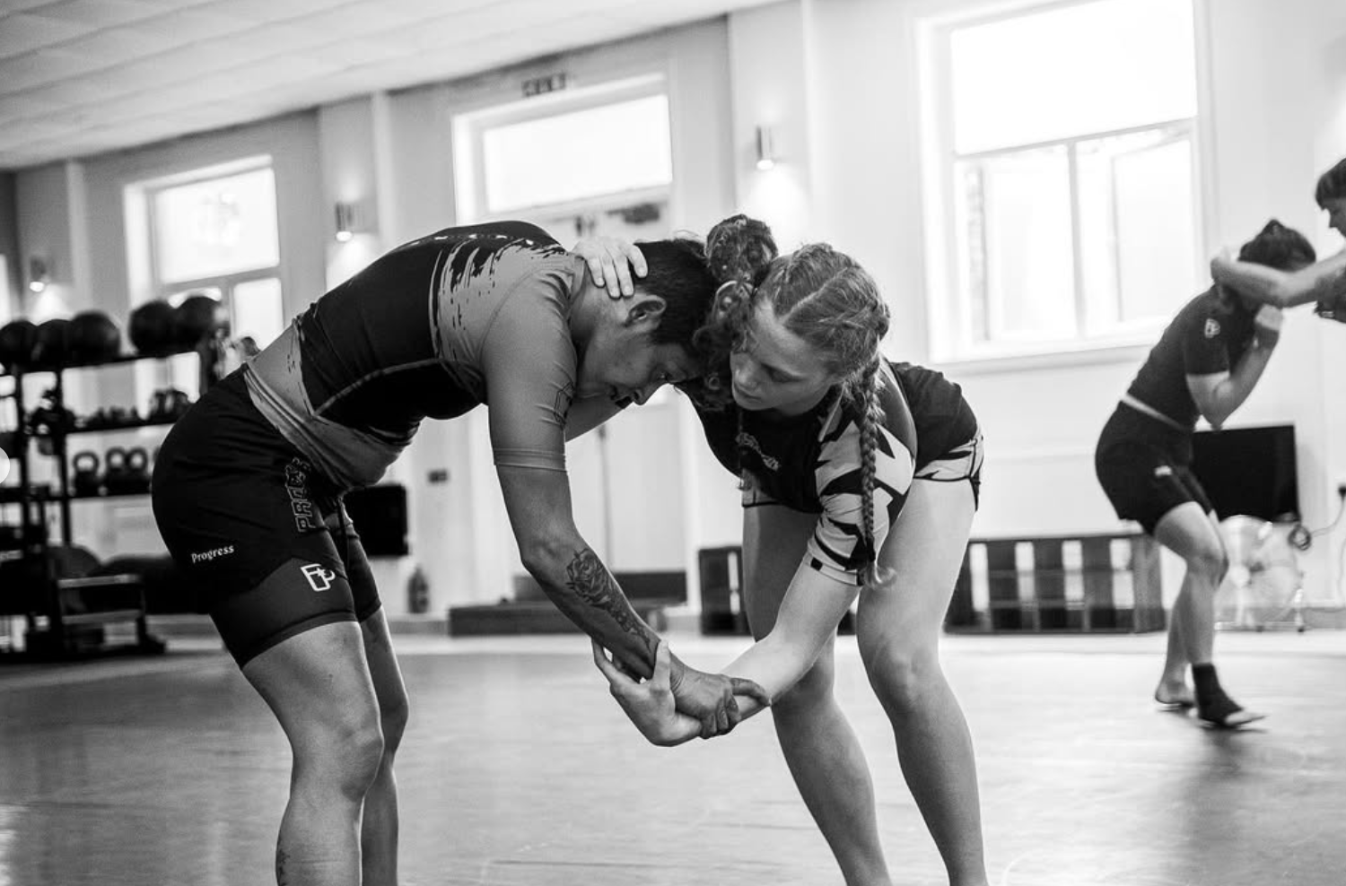 Two women practicing Brazilian jiu-jitsu at BJJ Academy SW18, one applying a submission hold on the other in a gym, with other practitioners in the background.