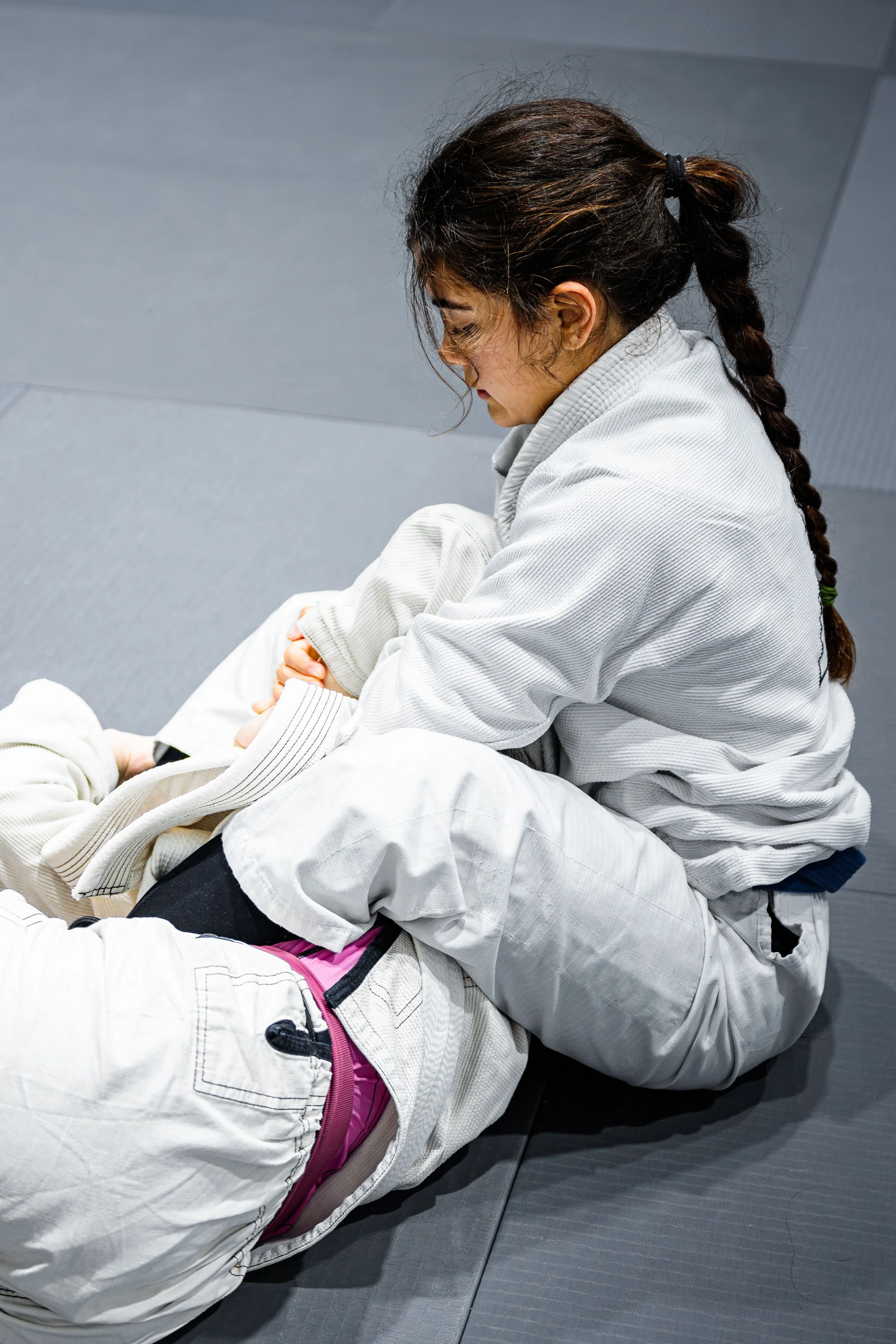 Two women practicing Brazilian Jiu-Jitsu in SW18 on a gray mat. One is wearing a white gi with pink accents, and the other is in a white gi with black hair tied back. The woman with darker hair is executing a grappling move.