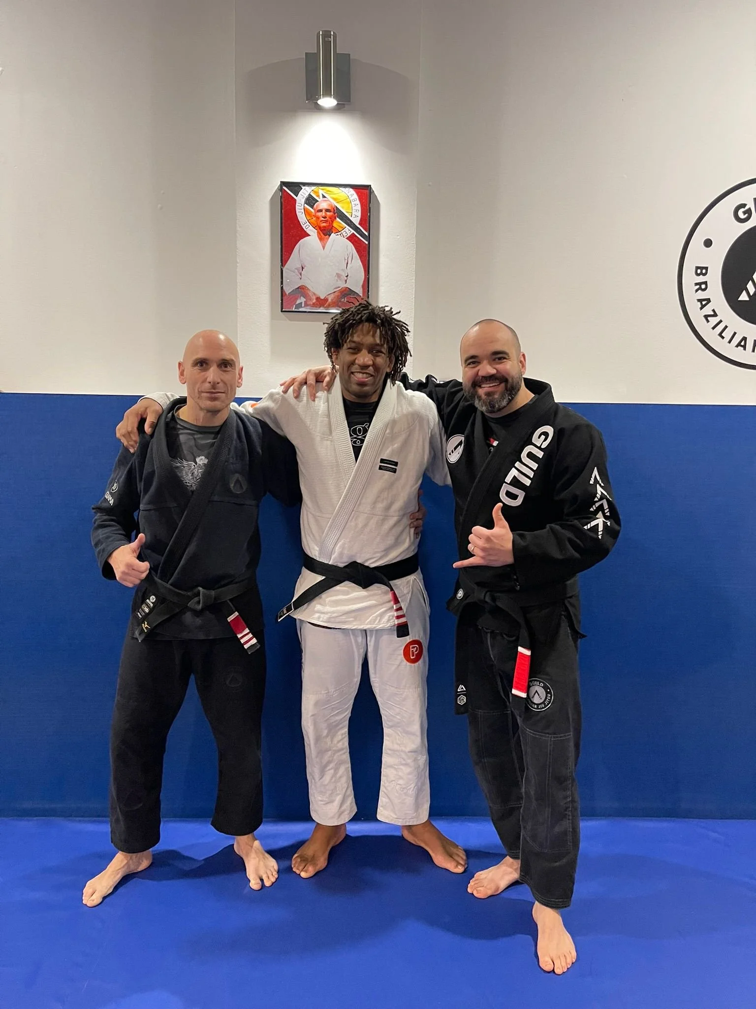 Three men in Brazilian Jiu-Jitsu gis at a Brazilian jiu jitsu academy in Wandsworth, London, posing together on a blue mat. One man is wearing a white gi, and the other two are wearing black gis. They are smiling.