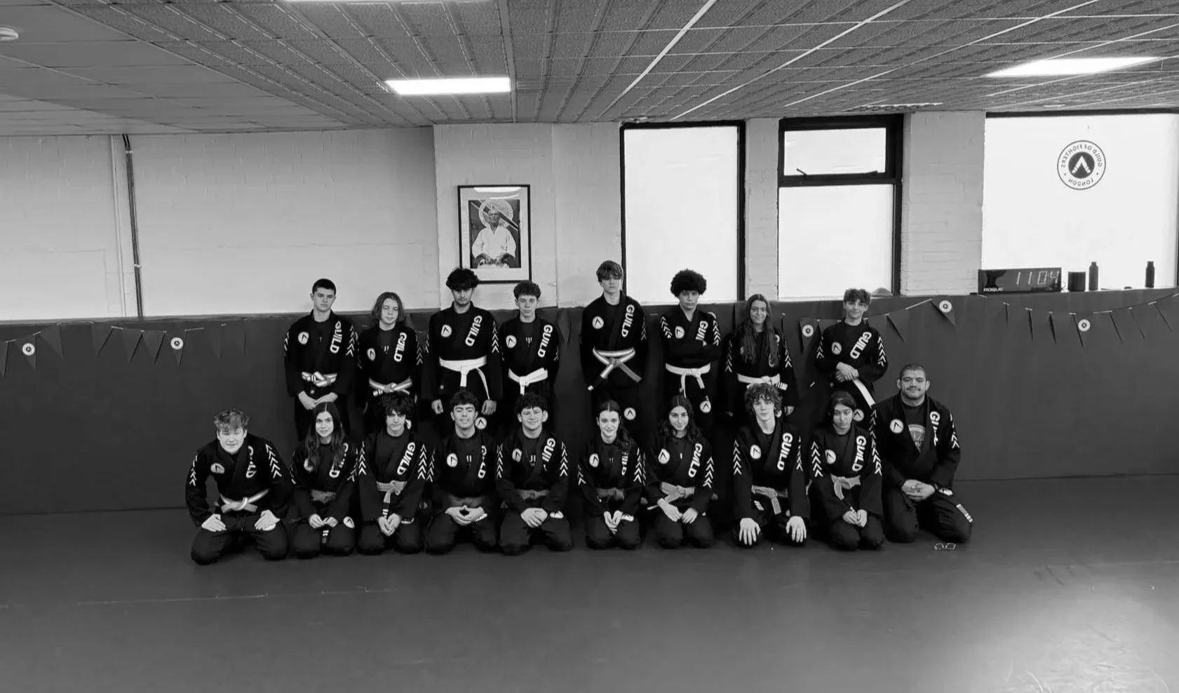 Group photo of teens BJJ class in South West London in black gi uniforms with white belts, posed in a gym with targets on the mat and a wall with a picture and target logo.