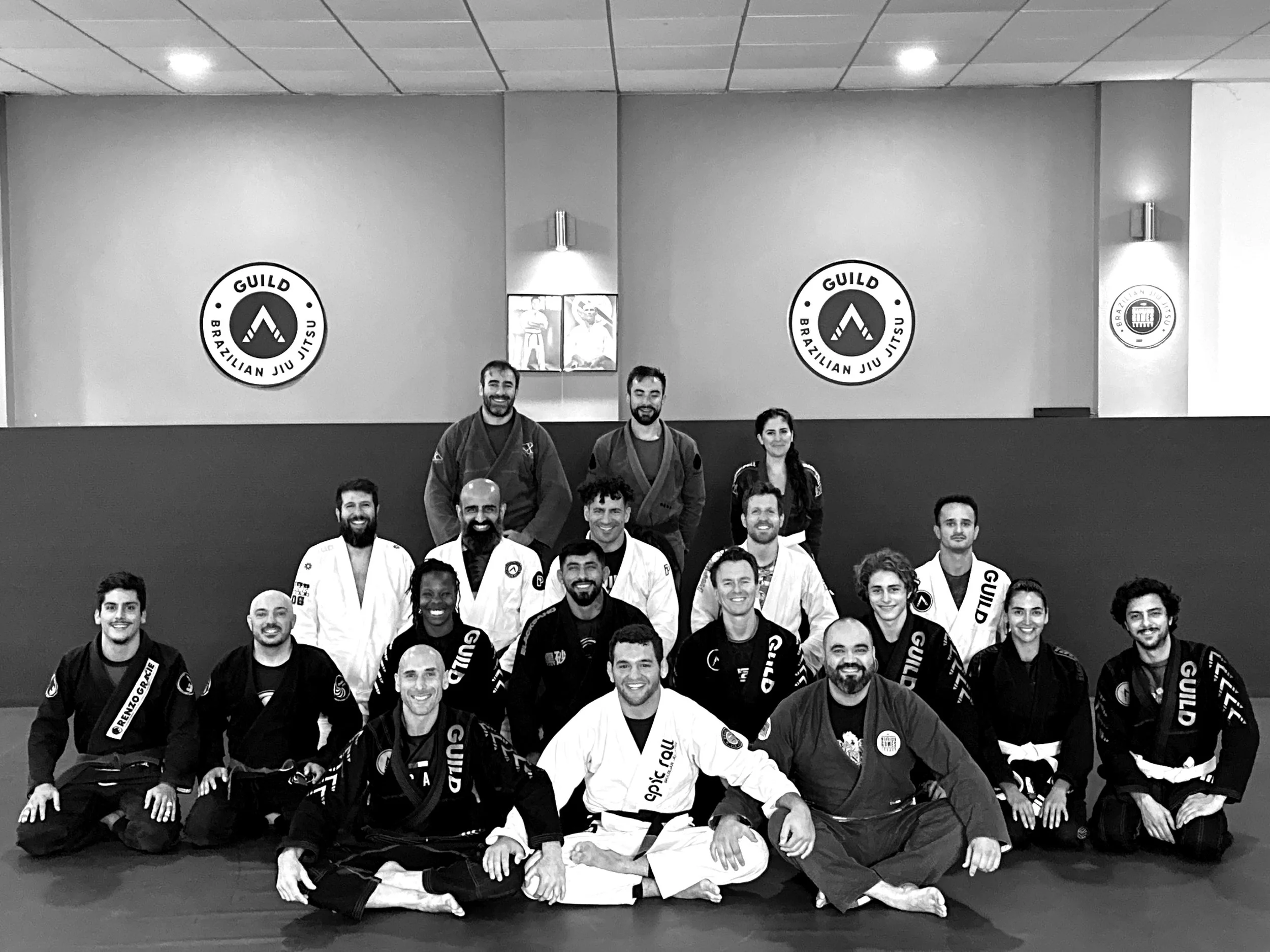 Group photo of Brazilian Jiu Jitsu practitioners in London in gis, standing together in a dojo, with visible mats and banners on the wall behind them.