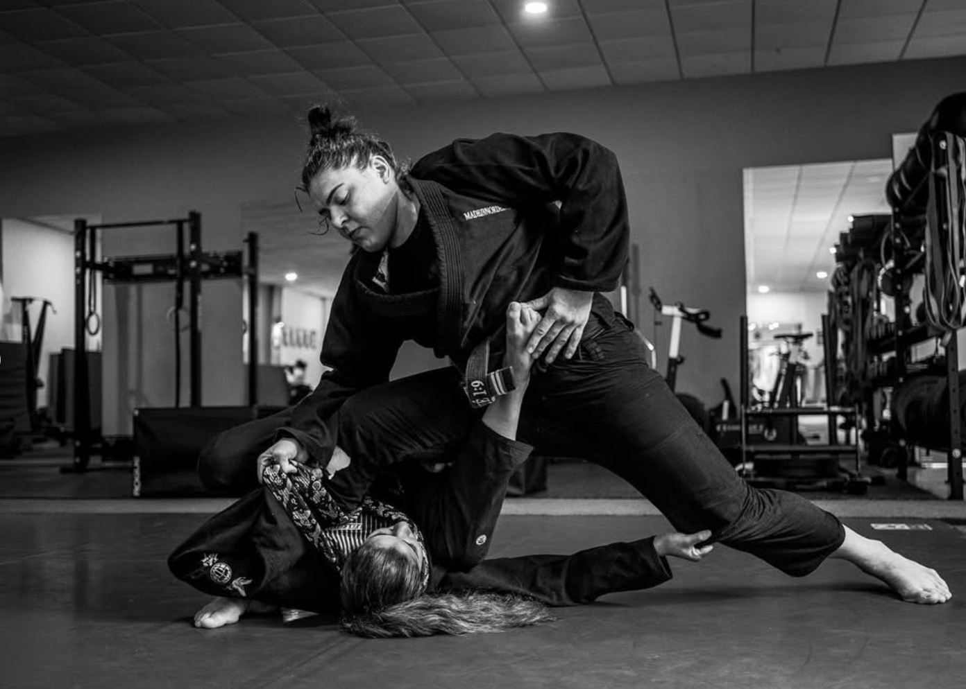 Two women practicing Brazilian Jiu-Jitsu on a mat in a gym. One woman is in a dominant position, applying a chokehold on the other woman, who is lying on the mat. Both are wearing gis.