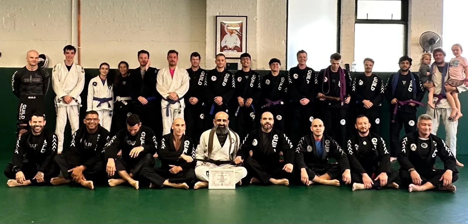 Group photo of Brazilian Jiu-Jitsu instructors in Wandsworth, London in gi uniforms, posing in a dojo with a green mat, a framed picture on the wall, and windows in the background.