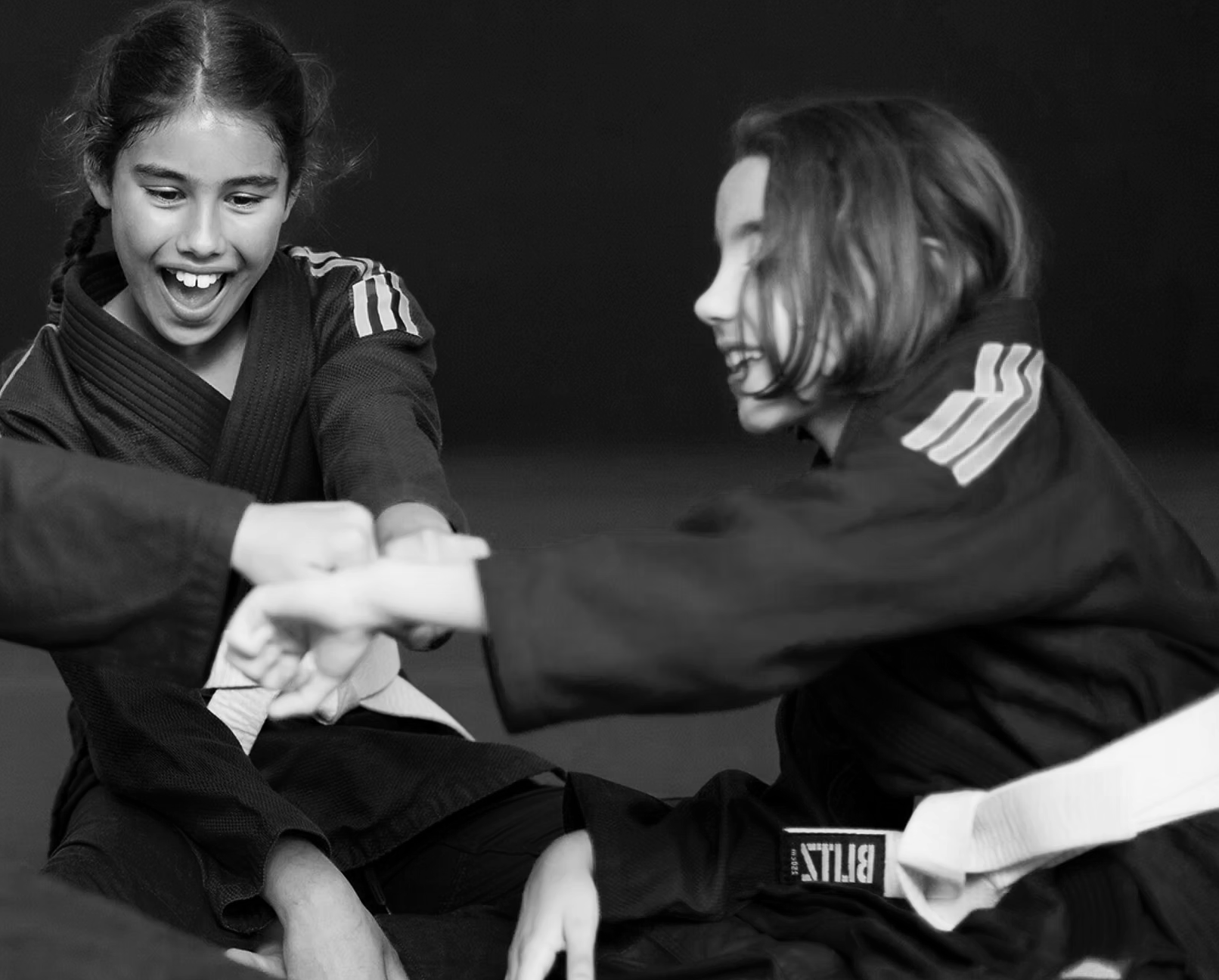 Two children dressed in martial arts Gis engaged in a judo match, smiling and holding each other's hands, against a dark background.