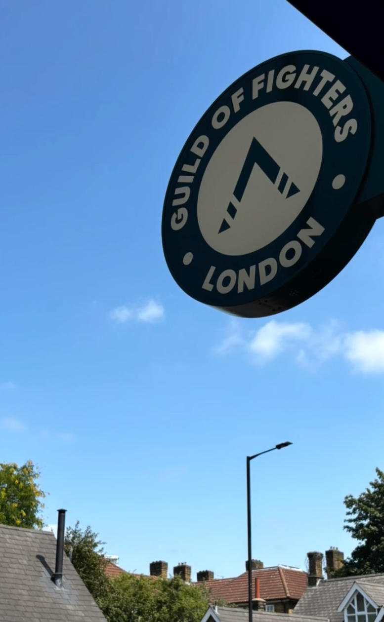 Close-up of a circular sign for the Guild of Fighters in London, a Brazilian jiu jitsu academy in south west London, featuring a stylized fighter logo against a blue sky, with rooftops and street lamps in the background.