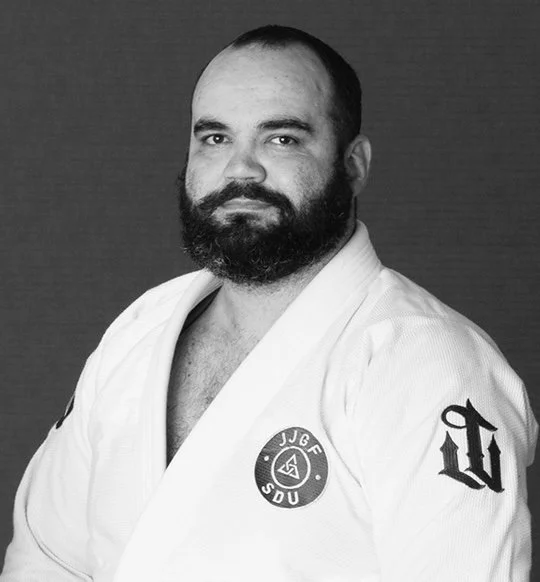 Man with a beard wearing a martial arts uniform with patches, looking at the camera.