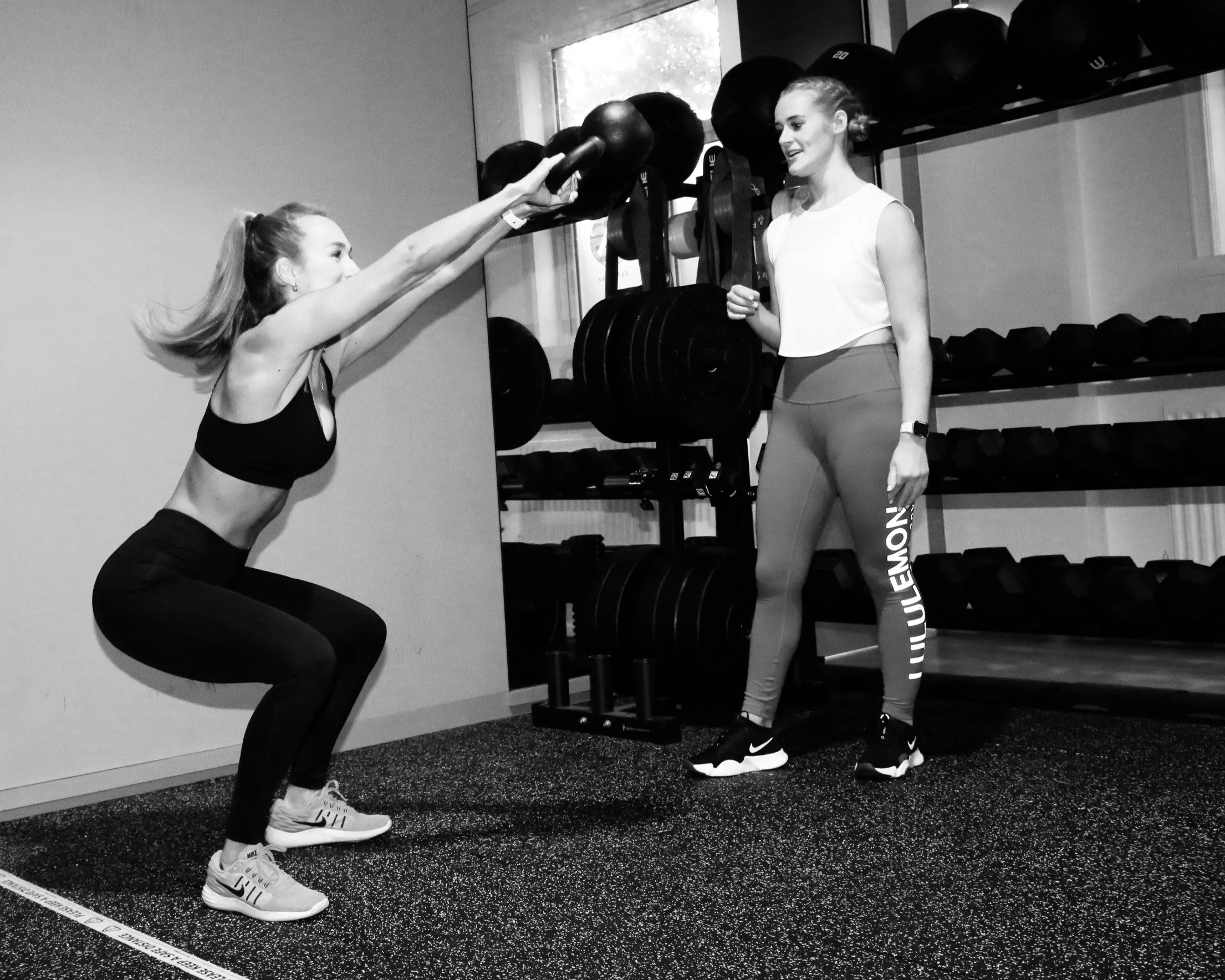 A woman with long hair tied back, wearing a sports bra and leggings, is squatting and holding a medicine ball with both hands. She is in a gym, facing another woman who is standing, wearing a sleeveless top and leggings, and is smiling. The standing 