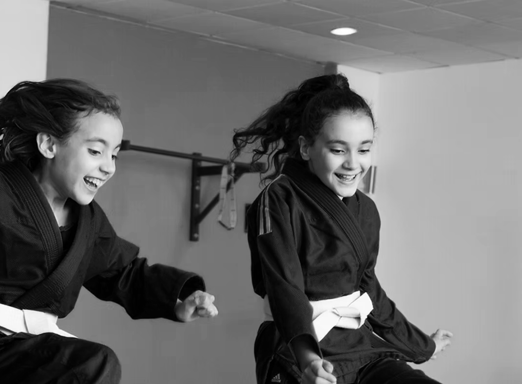 Two young girls smiling and wearing black martial arts uniforms with white belts, practicing martial arts moves together in a dojo.
