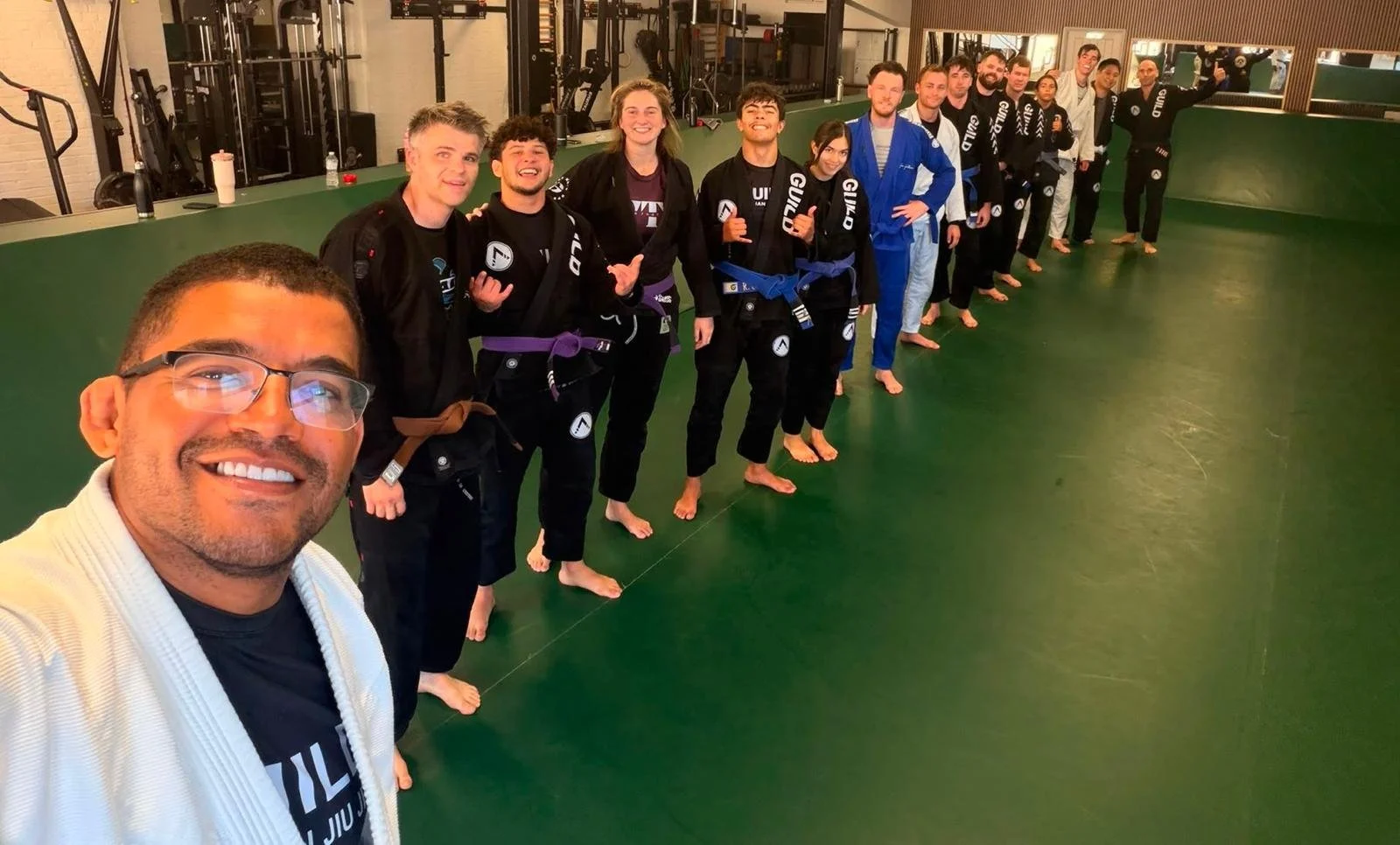 A group of people in Brazilian Jiu-Jitsu gis standing in a line on a green mat, inside a martial arts gym, taking a group selfie. The person in the foreground is a man with glasses, smiling at the camera, wearing a white gi and black shirt. The other individuals are also smiling and some are giving thumbs-up. The gym has various equipment visible in the background.