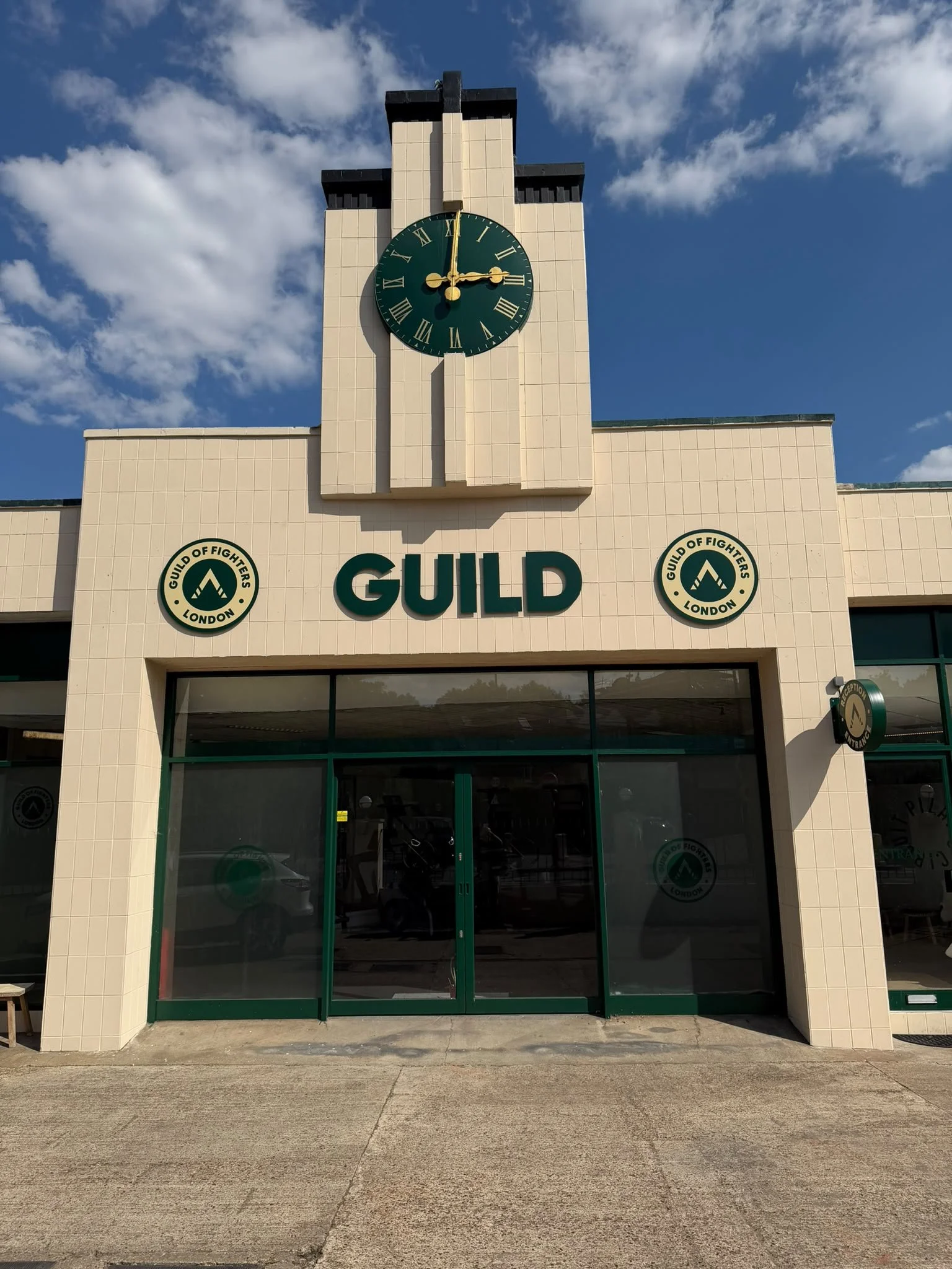 Exterior of the jiu-jitsu academy SW18 London building with a clock showing 3:00, the sign reads 'GUILD' and has the logo of the Guild of Fighters London, with a blue sky and scattered clouds in the background.