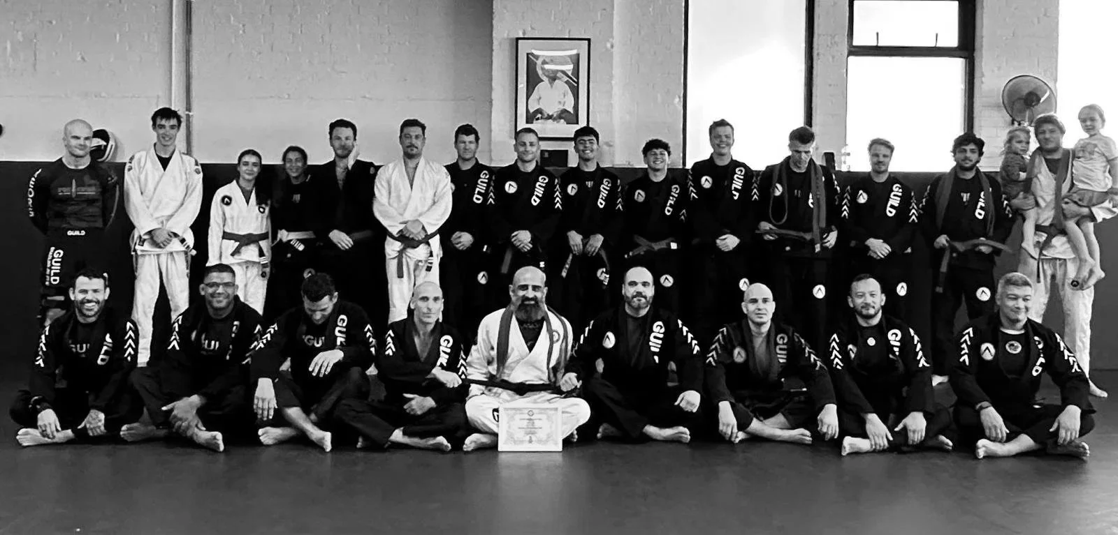 Group photo of martial arts practitioners in Wandsworth, London, some in white gi, others in black gi, sitting and standing on a training mat in a dojo, with a framed photo and window in the background.