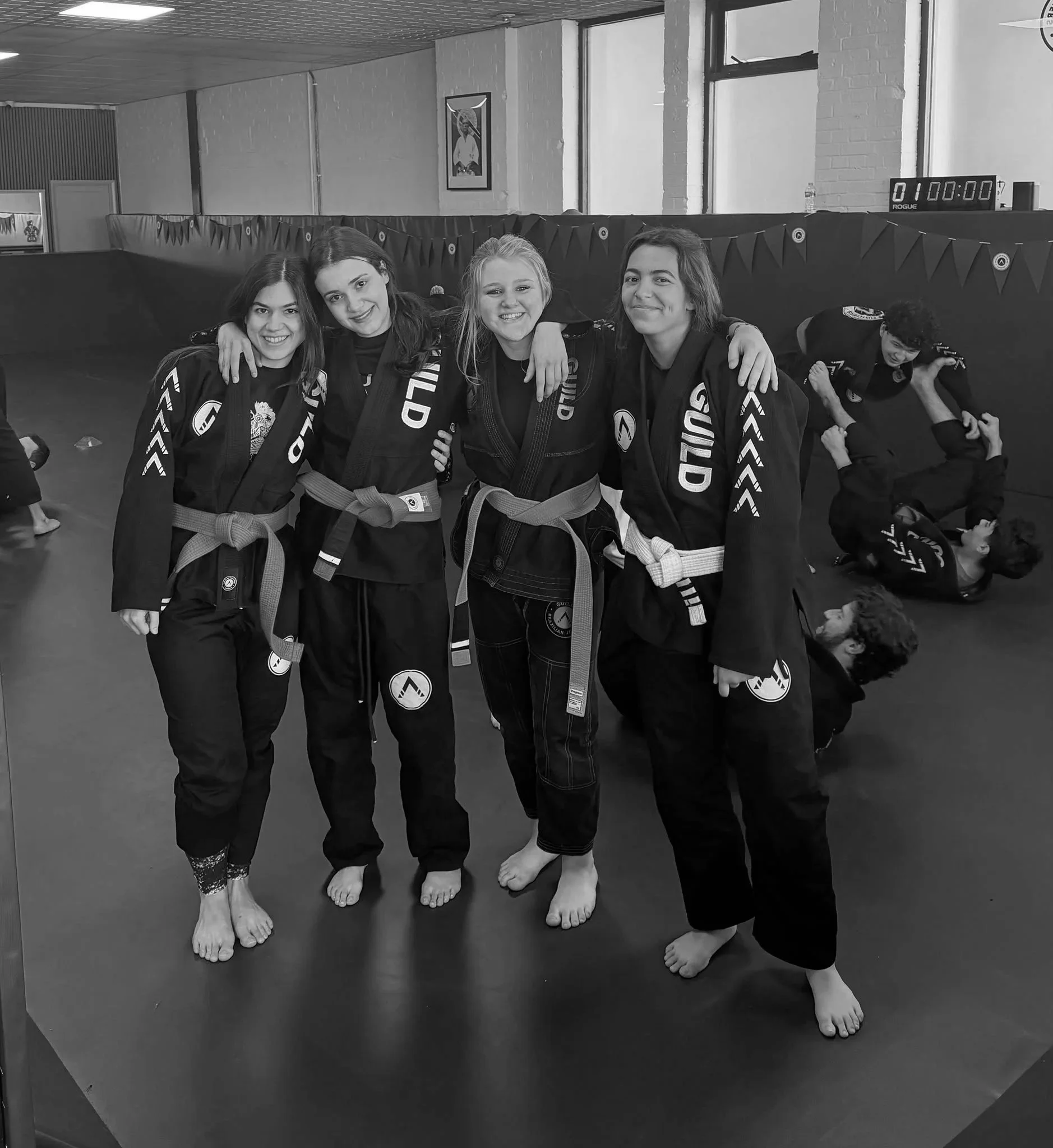 Four women in martial arts uniforms standing together with arms around each other, smiling, inside a martial arts training facility.