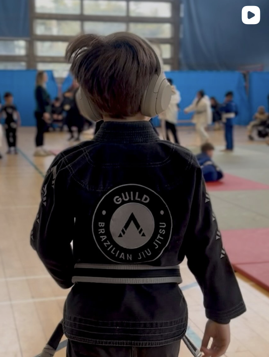 Child wearing headphones and a Brazilian Jiu Jitsu gi with a guild logo on the back, standing in a gymnasium filled with people and martial arts mats.