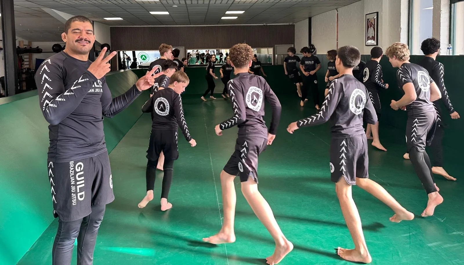 Martial arts class in a gym with children and an instructor, all wearing matching black uniforms with white logo and text, practicing Techniques on a green mat.